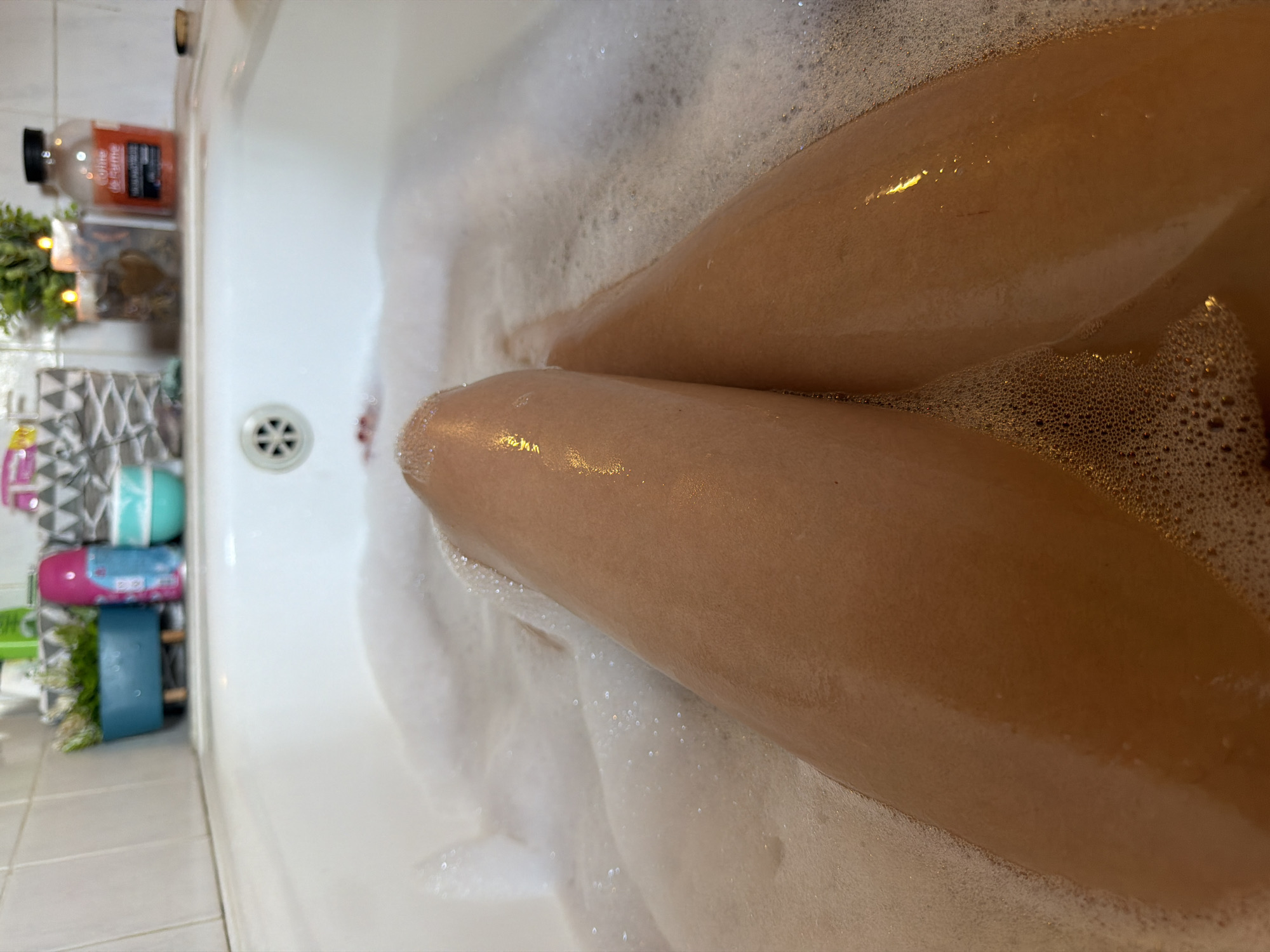 The media file shows a close-up view of a person's legs submerged in a bubbly bathtub. The legs, with smooth skin reflecting light, are the central focus. The scene is a bathroom, indicated by the white bathtub and tiled floor. Various bath products, including bottles and what appear to be decorative items, are visible on the edge of the tub to the left. The time of day and weather are not discernible. The overall impression is one of relaxation and self-care. There is no visible text in the media file.