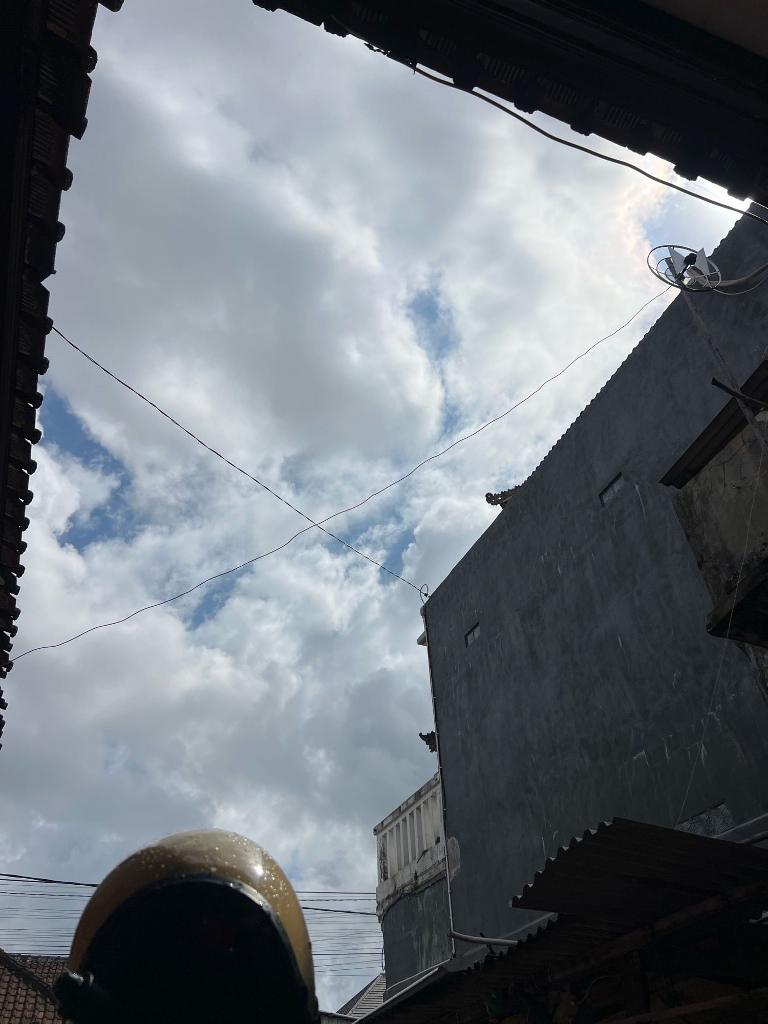 A low-angle shot captures a cloudy sky framed by dark, horizontal roof structures. In the foreground, the rounded, golden surface of a helmet is visible, its surface dotted with water droplets. The mid-ground features a tall, dark gray building with a corrugated metal roof and a small satellite dish mounted on its upper wall. Two thin wires cross the sky diagonally. The background is dominated by a sky filled with white, billowy clouds interspersed with patches of blue. The scene appears to be an urban environment, possibly in Denpasar, Indonesia, judging by the architectural style and atmospheric conditions.