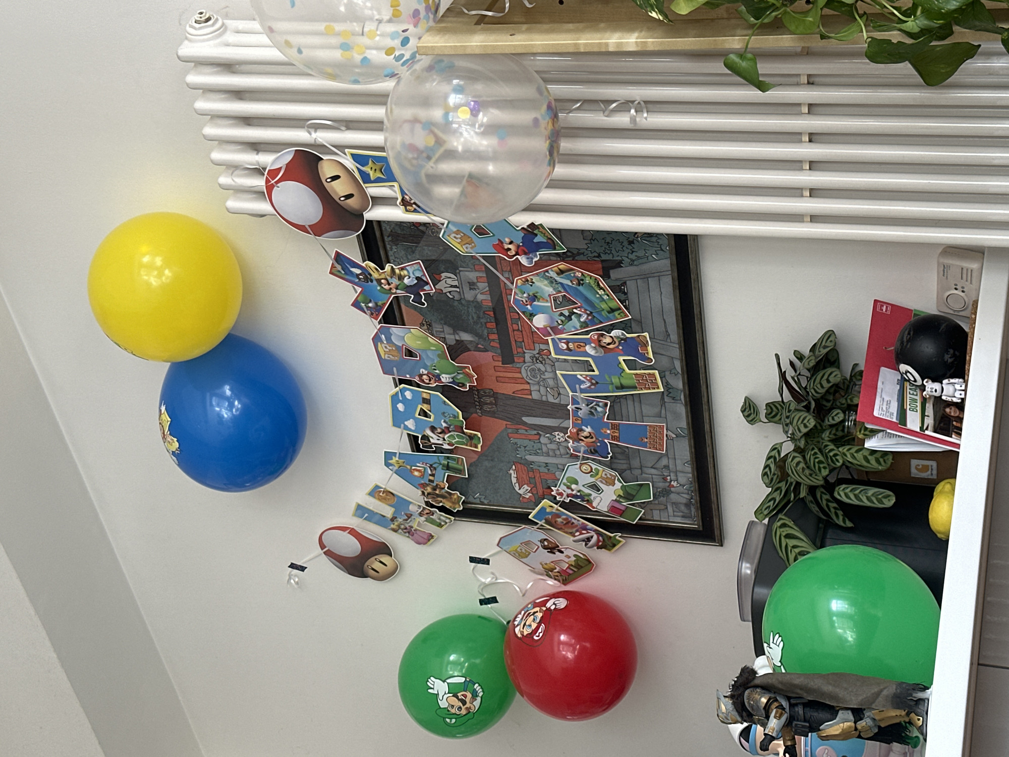 The image depicts a celebratory scene indoors, likely for a birthday, given the presence of balloons and a banner spelling out "HAPPY 4TH BIRTHDAY" adorned with Super Mario characters. The balloons are in a range of colors, including yellow, blue, green, and red, some with Mario-themed decorations. A clear balloon filled with confetti is also visible. The decorations are affixed to a white wall, with a white radiator behind them, and a framed Super Mario poster. To the right of the framed poster, there's a collection of objects including a plant, a yellow lemon, and what appears to be a figure. Given that no information in the image indicates location in London, United Kingdom, the location details cannot be definitively ascertained.
