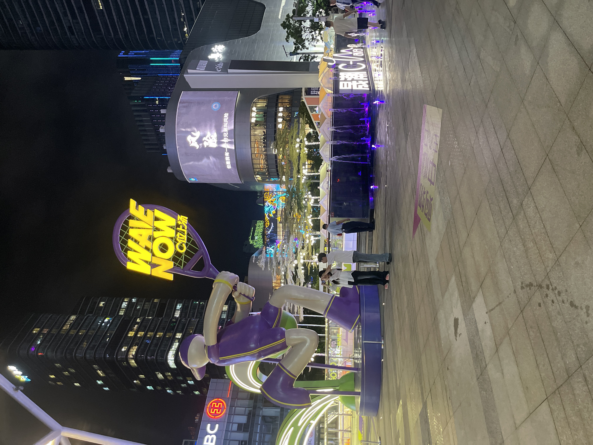 This media file, captured in Panyu District, China, depicts a lively nighttime scene in an urban plaza. The primary subjects are a large, colorful sculpture of a tennis player holding a racket, illuminated with neon lights. The sculpture is vibrant, with a purple outfit and yellow accents, and features the text "WAVE NOW" and "C位上场" (meaning "enter at the C position" or "play like a star"). The scene is set against a backdrop of modern, tall buildings with illuminated windows, suggesting a city environment. In the middle ground, a fountain with purple lighting adds to the ambiance, with several people standing around it. The ground is paved with large square tiles, some of which appear wet, possibly from the fountain or recent rain. The overall mood is festive and energetic, with the bright lights and urban setting contributing to a dynamic atmosphere. The time of day is clearly night, indicated by the darkness of the sky and the prominent artificial lighting. Visible text also includes "BC E3" on a sign below the sculpture and "风险" (meaning "risk") on a screen on the building.