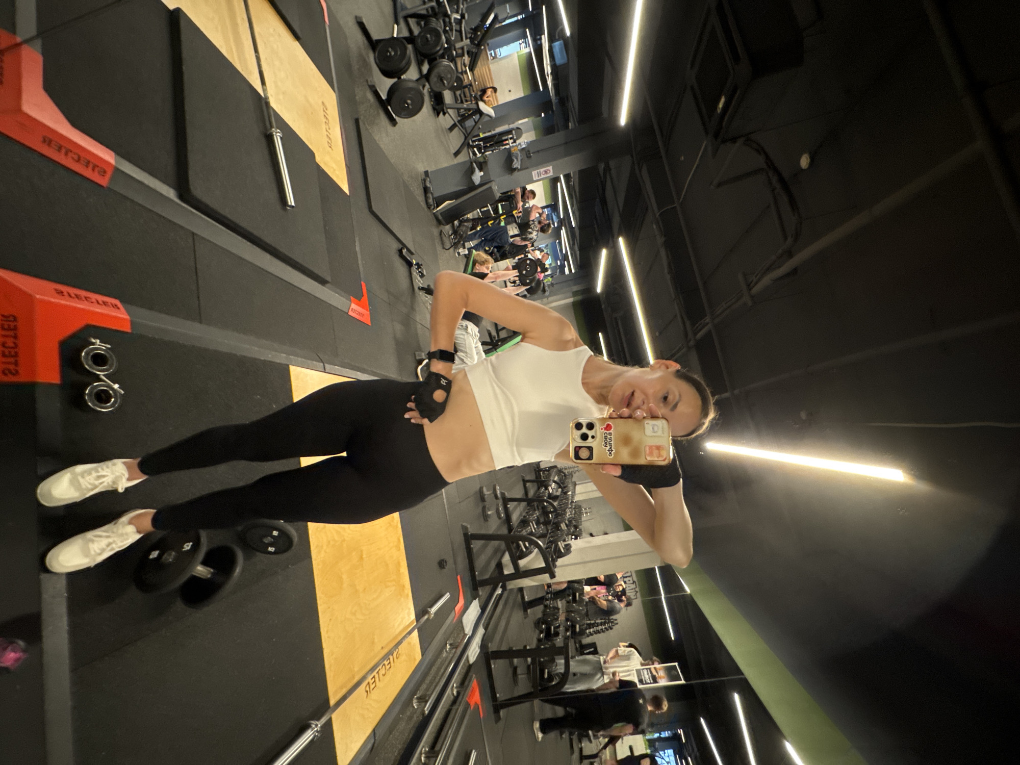 A woman in athletic wear is taking a mirror selfie in a gym. She is wearing a white tank top, black leggings, and white sneakers. She has on fingerless gloves. The gym appears to be well-equipped with various weight machines, dumbbells, and weight plates visible in the background. Other people are also working out in the gym, some visible in the reflections of the mirrors. The gym has dark walls and a black ceiling with strip lighting. The floor is a combination of black rubber mats and wooden platforms. On the woman's phone case, there is visible text. The overall atmosphere appears to be energetic and focused on fitness. The lighting suggests it could be daytime or evening, as the artificial lights are on. The location cue provided is Sochi, Russia.