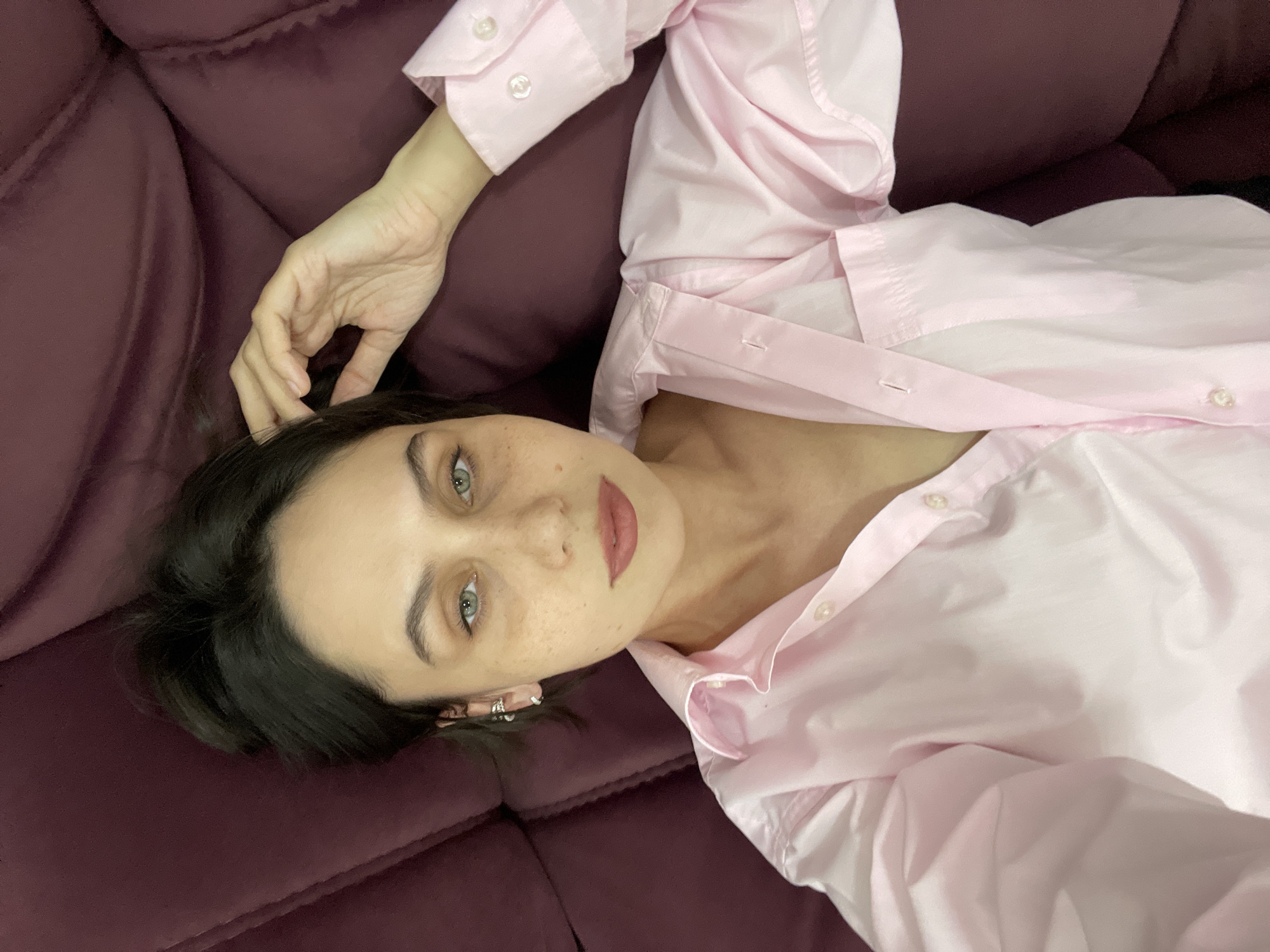 A woman with fair skin, dark hair, and green eyes is lying on a dark maroon, possibly velvet, couch. She is wearing a light pink button-down shirt, partially unbuttoned. Her right hand is near her face, touching her hair. She has freckles on her face and is wearing multiple earrings in her left ear. The photograph appears to be taken from an overhead perspective. The location is not apparent from this image.