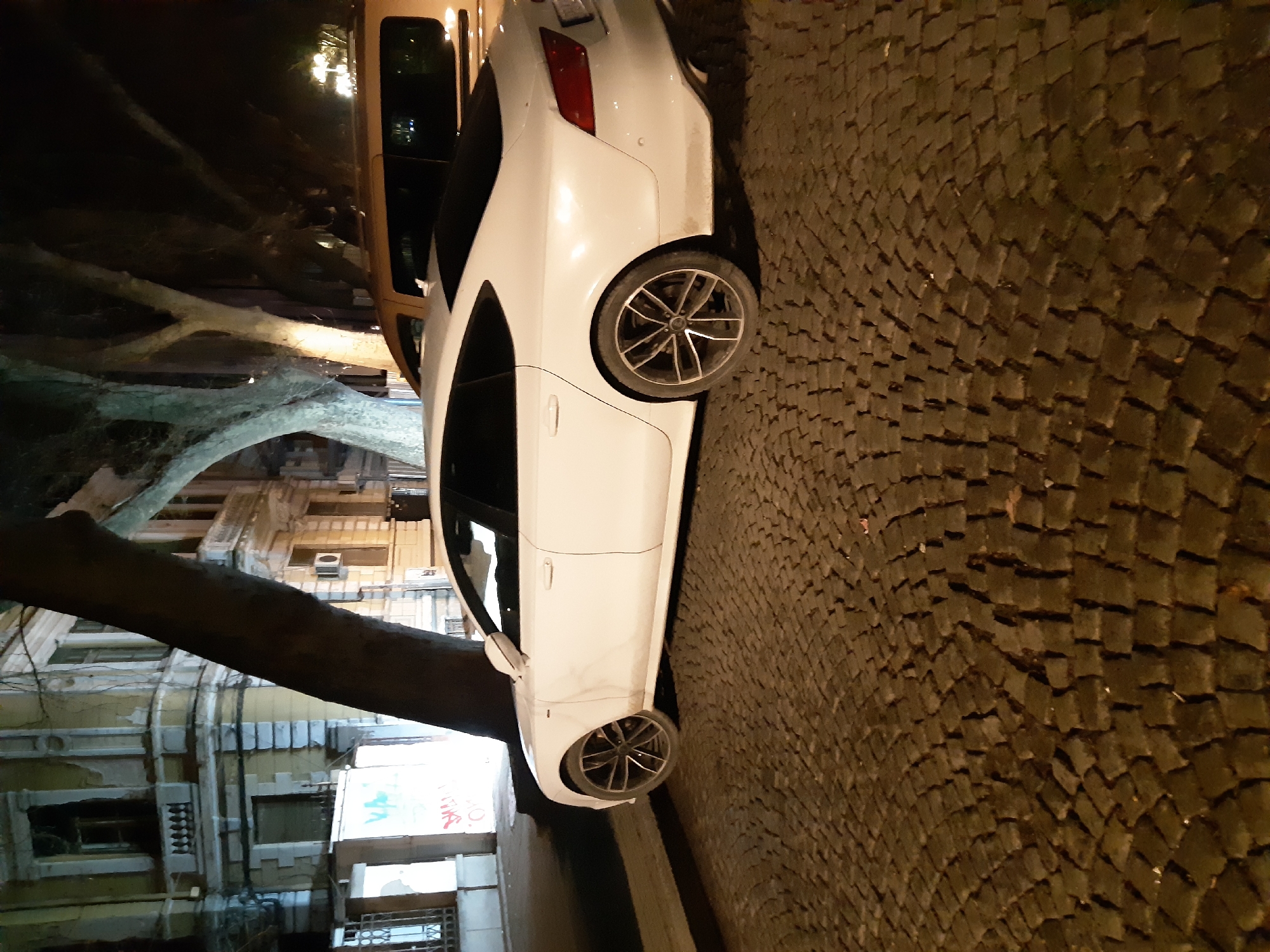 A white car is parked on a cobblestone street in Plovdiv, Bulgaria. The car is partially obscured by a tree branch on the left. The background features a building with classical architectural details. The scene is illuminated by streetlights.