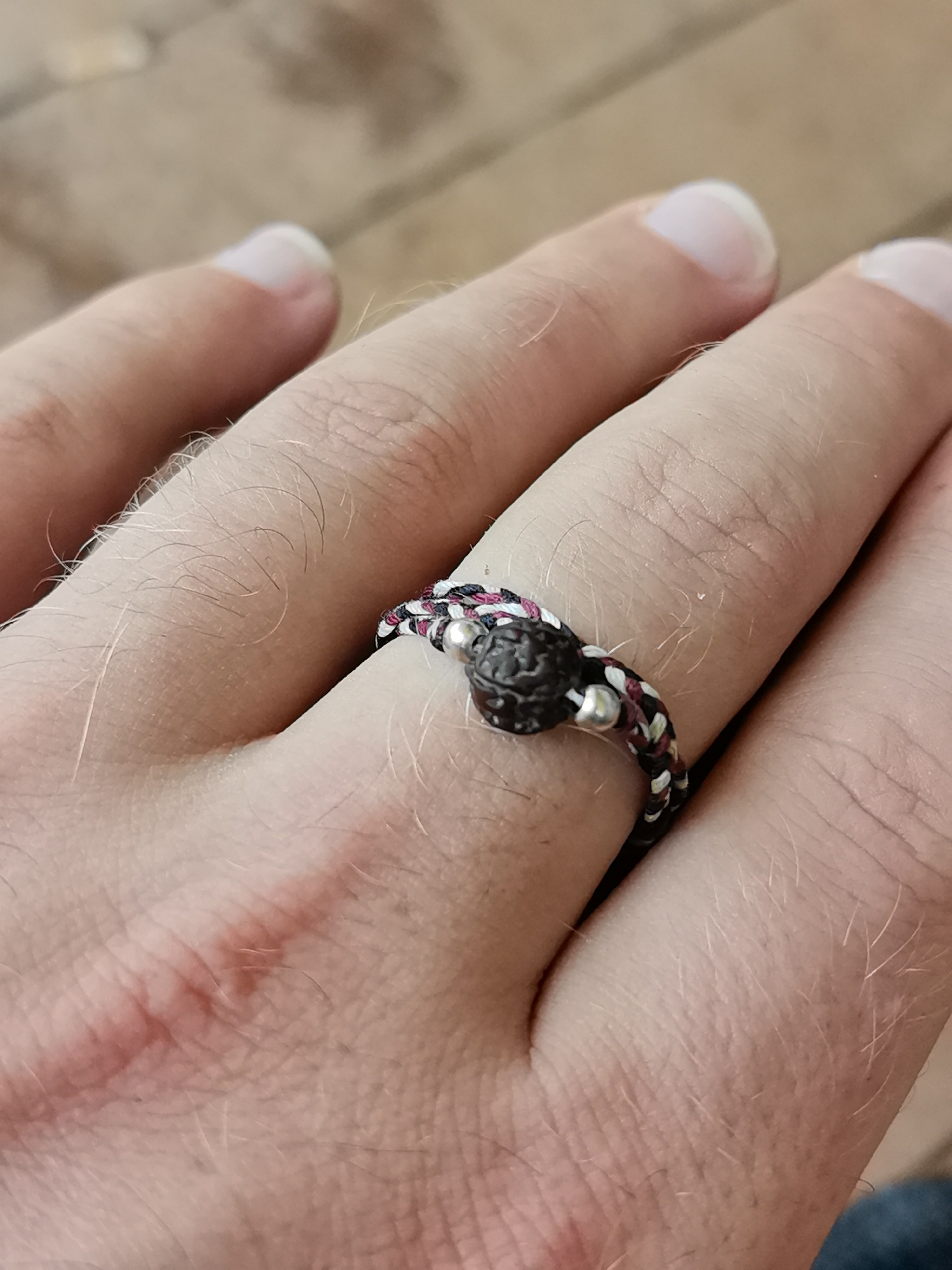 A close-up, eye-level shot shows a person's hand, finger, and a ring. The ring is made of braided cords in black, white, and maroon, with a dark, textured bead as the centerpiece and two small silver beads on either side. The ring is worn on the middle finger of the left hand, which rests on a surface that appears to be made of wood planks, suggested by the blurred background. The skin on the hand is fair and has visible hairs. There is a faint reddish mark on the palm side of the hand, possibly a scar or an abrasion. The lighting is natural and casts soft shadows. The image is in focus on the ring and the finger. The specific location within Bac Nha Trang Ward, Vietnam, cannot be determined from the visual information provided in the image.