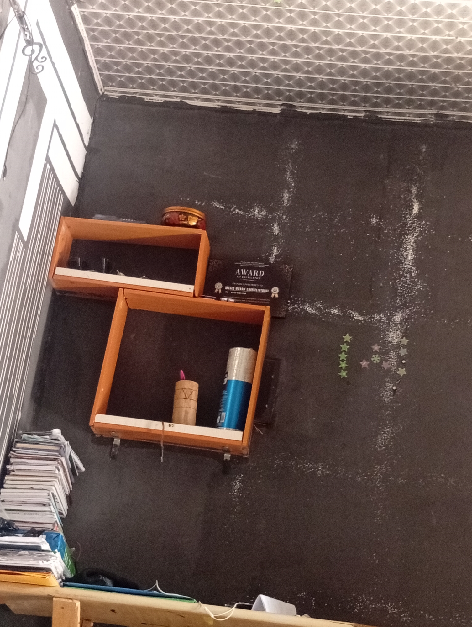 The image depicts a corner of a room with dark grey walls and a white patterned ceiling. On the left side, there are white and black diagonal stripes on the wall. Two wooden shelves are mounted on the dark grey wall. The top shelf holds a small, round, red and gold decorative tin. The bottom shelf contains a cylindrical object, possibly a candle, with a pink wick and some markings, and a blue and silver cylindrical object, likely a can of spray paint. To the left of the shelves, a stack of books and papers is piled on a wooden surface. On the wall above and to the right of the shelves, there is a black plaque with gold lettering that reads "AWARD OF EXCELLENCE" and further text. To the right of the plaque, a cluster of small, colorful star-shaped stickers are affixed to the wall. The wall itself appears dusty or speckled with white material. The overall impression is of a decorated interior space, possibly a home office or study area.