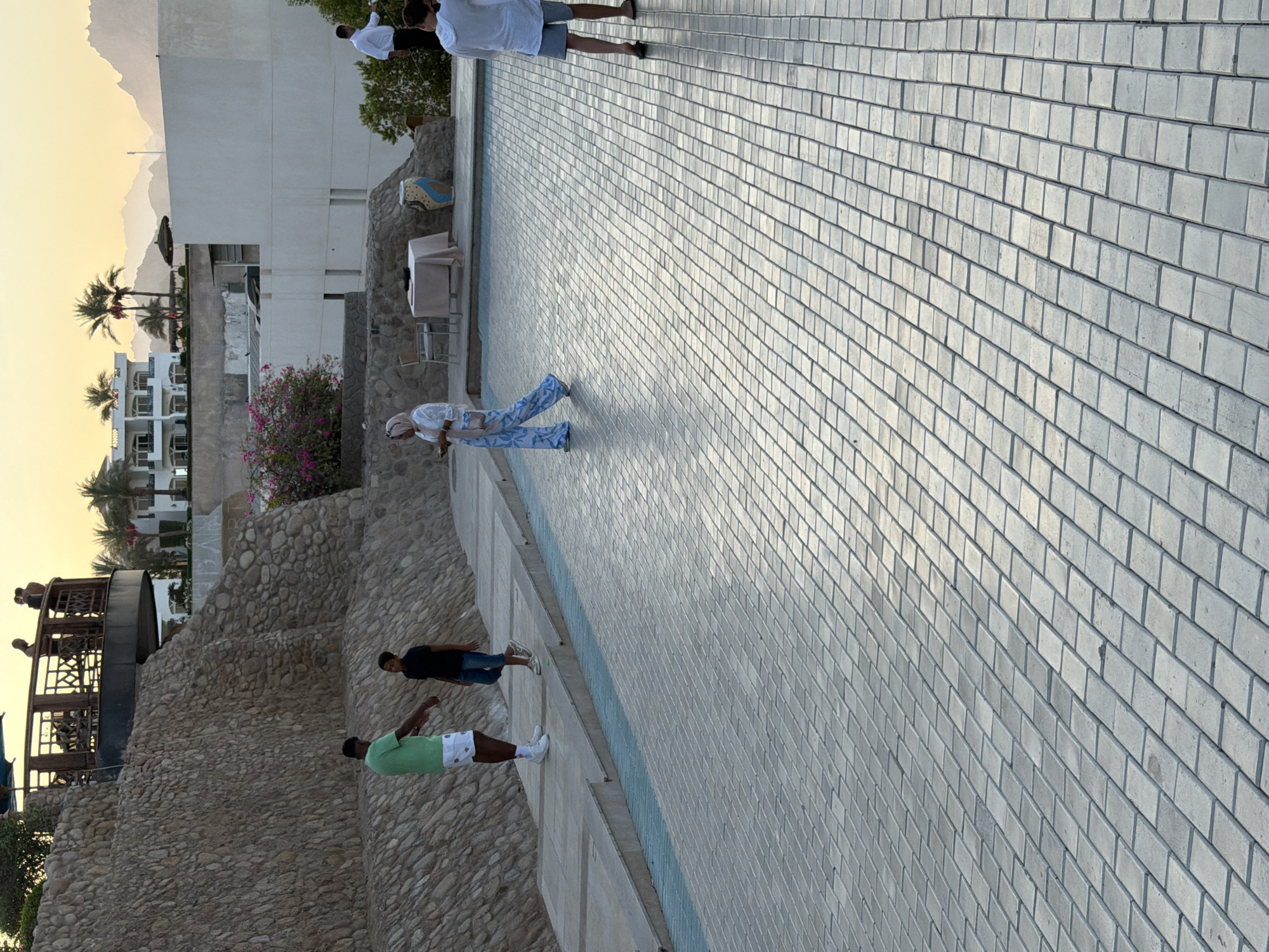 The image depicts an outdoor scene with a paved area and a stone wall in Sharm El Sheikh, Egypt. In the foreground, a tiled surface stretches into the distance. To the left, a man in a green t-shirt and white shorts stands near a young boy in a black t-shirt and blue jeans. Further back, a woman wearing a headscarf and patterned pants walks across the paved area. Behind them, a table is set with a tablecloth, suggesting an outdoor dining or seating area. In the background, buildings and palm trees are visible against a hazy sky, with mountains in the far distance. The overall setting appears to be a resort or public space.