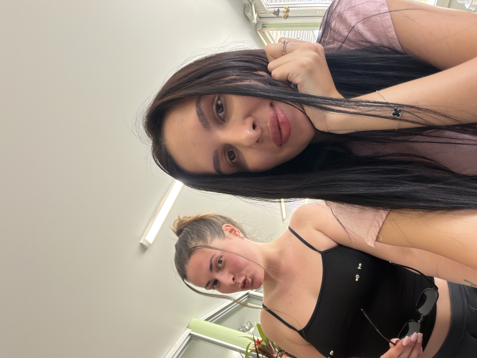 Two women are posing for a selfie indoors, likely at home or an office. The woman on the right is in the foreground, her face filling most of the frame. She has long, dark hair and is wearing a pink t-shirt. Her right hand is resting on her chin, and she is wearing rings on her fingers and a bracelet on her wrist. The woman on the left is in the background, slightly out of focus. She has her hair pulled up in a bun and is wearing a black tank top with silver embellishments. She is holding a pair of sunglasses in her left hand and has a playful expression on her face. The background appears to be a well-lit room with large windows and fluorescent lighting.