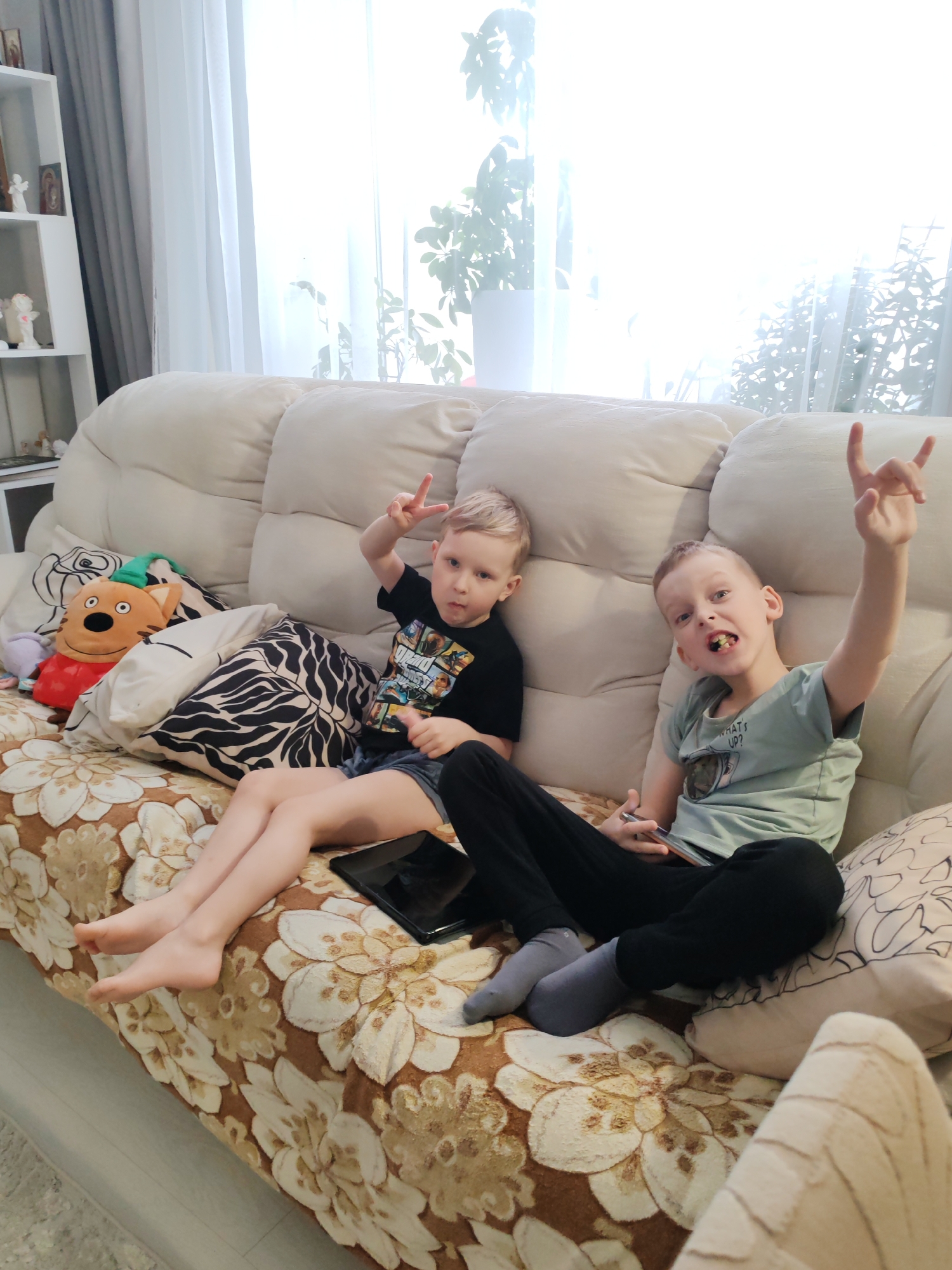 Two Caucasian boys are sitting on a beige floral patterned couch in a room with a white bookshelf and curtains. One boy is wearing a black t-shirt and denim shorts, holding a tablet and making a peace sign. The other boy wears a green t-shirt and black pants with socks, raising his hand in a "rock on" gesture. The location is inferred to be Chaikovsky, Russia. Pillows with different patterns are behind the boys, and a stuffed tiger toy is on the left side of the couch.