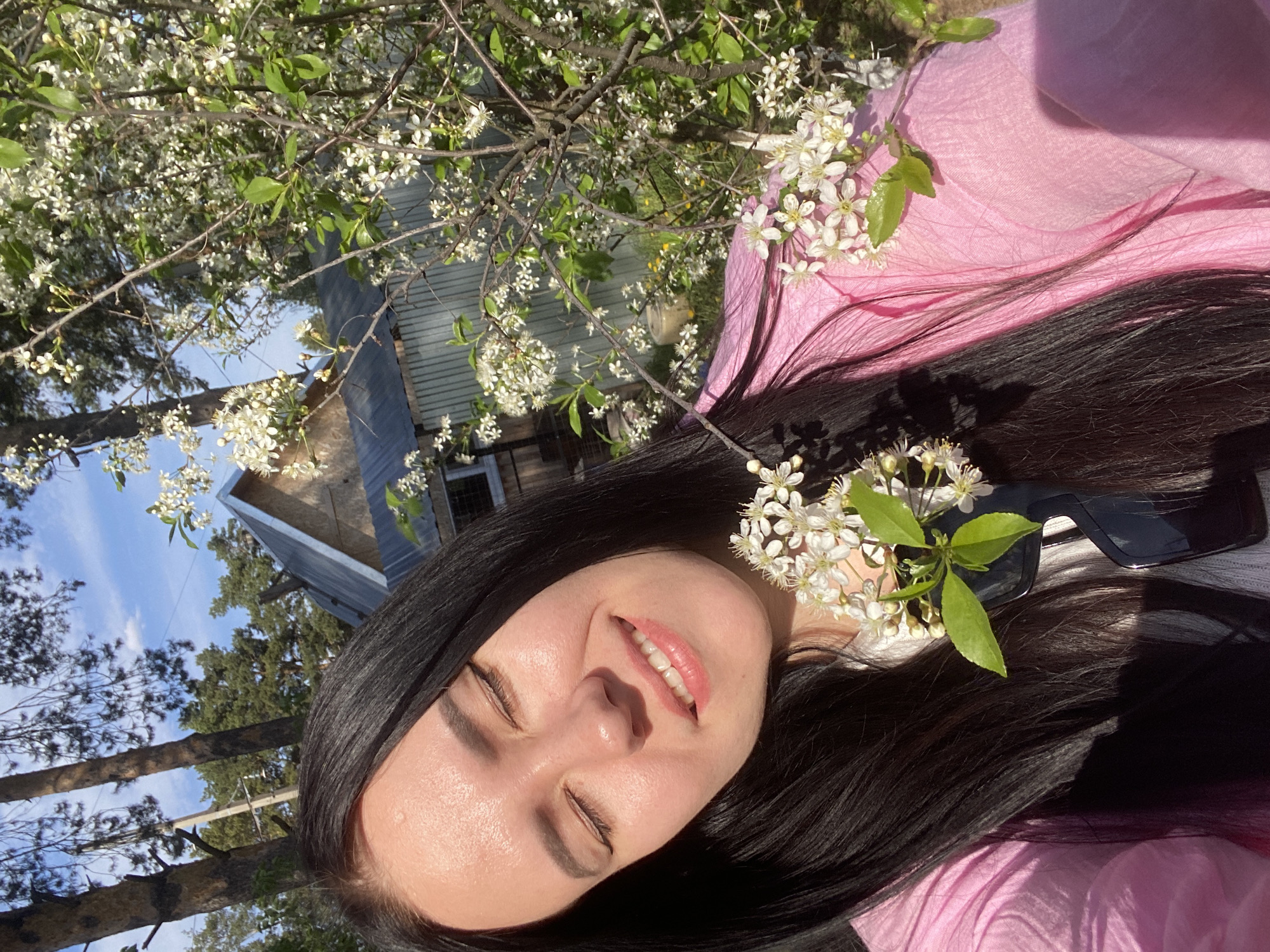 A low-angle, eye-level shot depicts a woman with long, dark hair, wearing a pink top and sunglasses, smiling with her eyes closed. She is positioned near a tree with white flowers. The background includes a wooden house and trees under a partly cloudy blue sky. The location is likely in a natural setting near the city of Chaikovsky, Russia. The focus is on the woman, with the flowers, house, and trees serving as background elements.