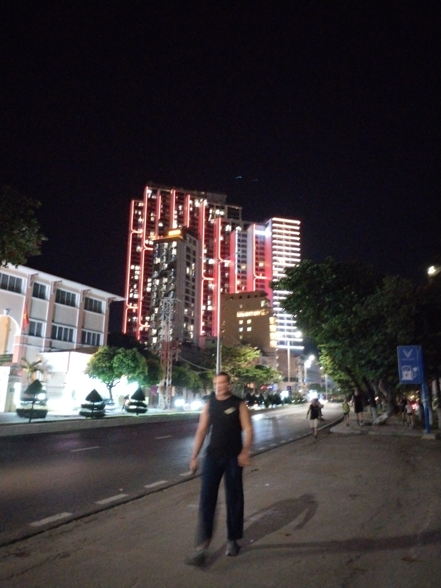 The image depicts a nighttime scene on a city street, likely in Bac Nha Trang Ward, Vietnam, as indicated by the presence of tall, brightly lit buildings which are characteristic of this area. In the foreground, a man in a dark sleeveless shirt and dark pants is walking towards the camera, his face blurred due to the low light conditions. He is casting a long shadow on the wet asphalt road.
The street is paved and appears to have been recently wet, reflecting the lights from the surrounding buildings and streetlights. White dashed lines mark the lanes of the road. On the left side of the image, there is a modern, light-colored building with multiple floors and windows. Adjacent to it and further down the street, several tall, high-rise buildings are illuminated with red and white lights, creating a vibrant urban nighttime atmosphere. Some trees and decorative bushes are planted along the sidewalk.
Further down the street, other individuals are visible, some walking and others appearing to stand. A blue sign with white icons is mounted on a pole near the sidewalk, possibly indicating a service or a restriction. The overall scene conveys a sense of urban activity at night.