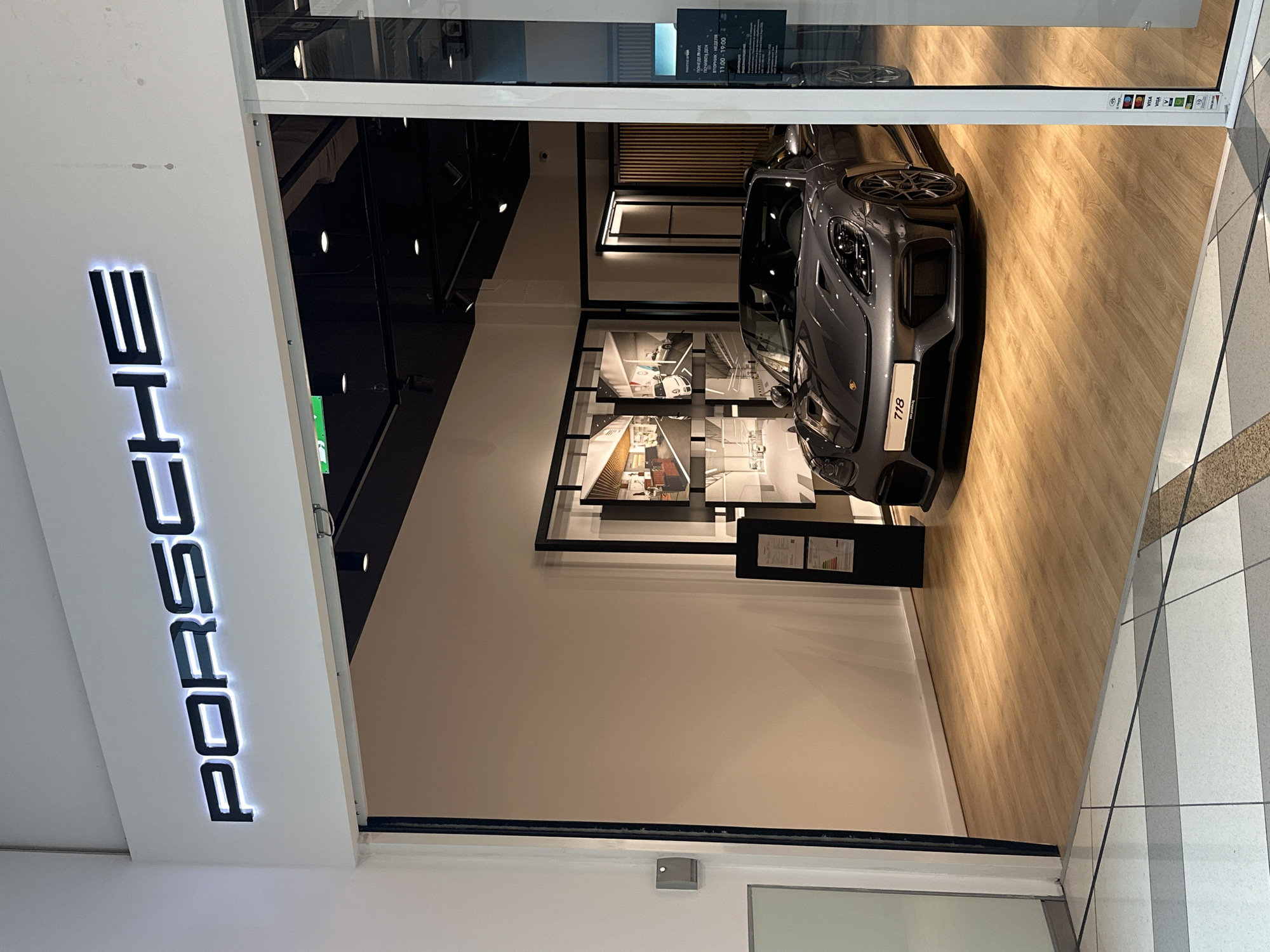 The image shows the exterior of a Porsche dealership in Varna, Bulgaria. The dealership has a modern facade with a prominent "PORSCHE" sign illuminated with white light. Inside, a gray Porsche 718 sports car is displayed on a wooden floor. The interior also features framed artwork on the walls and modern lighting fixtures. The dealership appears to be located within a shopping mall, as indicated by the tiled floor and the entrance area in the foreground. A sign on the glass door lists business hours and accepted payment methods.