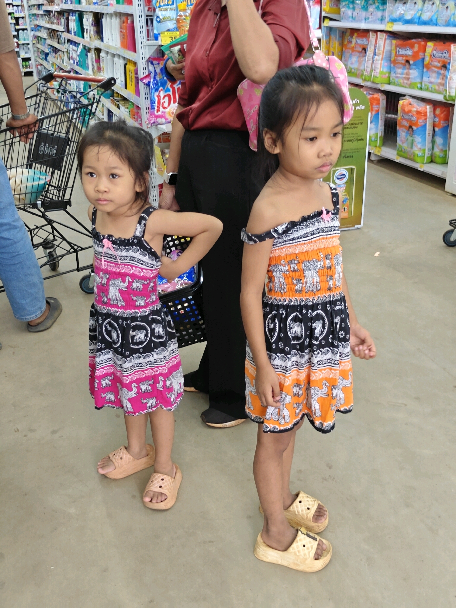 In Mu Si, Thailand, two young girls stand in a brightly lit aisle of what appears to be a grocery or department store. The girl on the left, wearing a black, pink, and white elephant-print dress and beige slides, looks towards the camera with a neutral expression. Next to her, the girl on the right, dressed in an orange, black, and white elephant-print dress and beige slides, looks to her left. Behind them, a shopping cart is partially visible, and shelves stocked with various products line the background. The floor is a light-colored, slightly textured material, and a person in dark pants and a maroon shirt is partially visible in the upper right quadrant of the image.