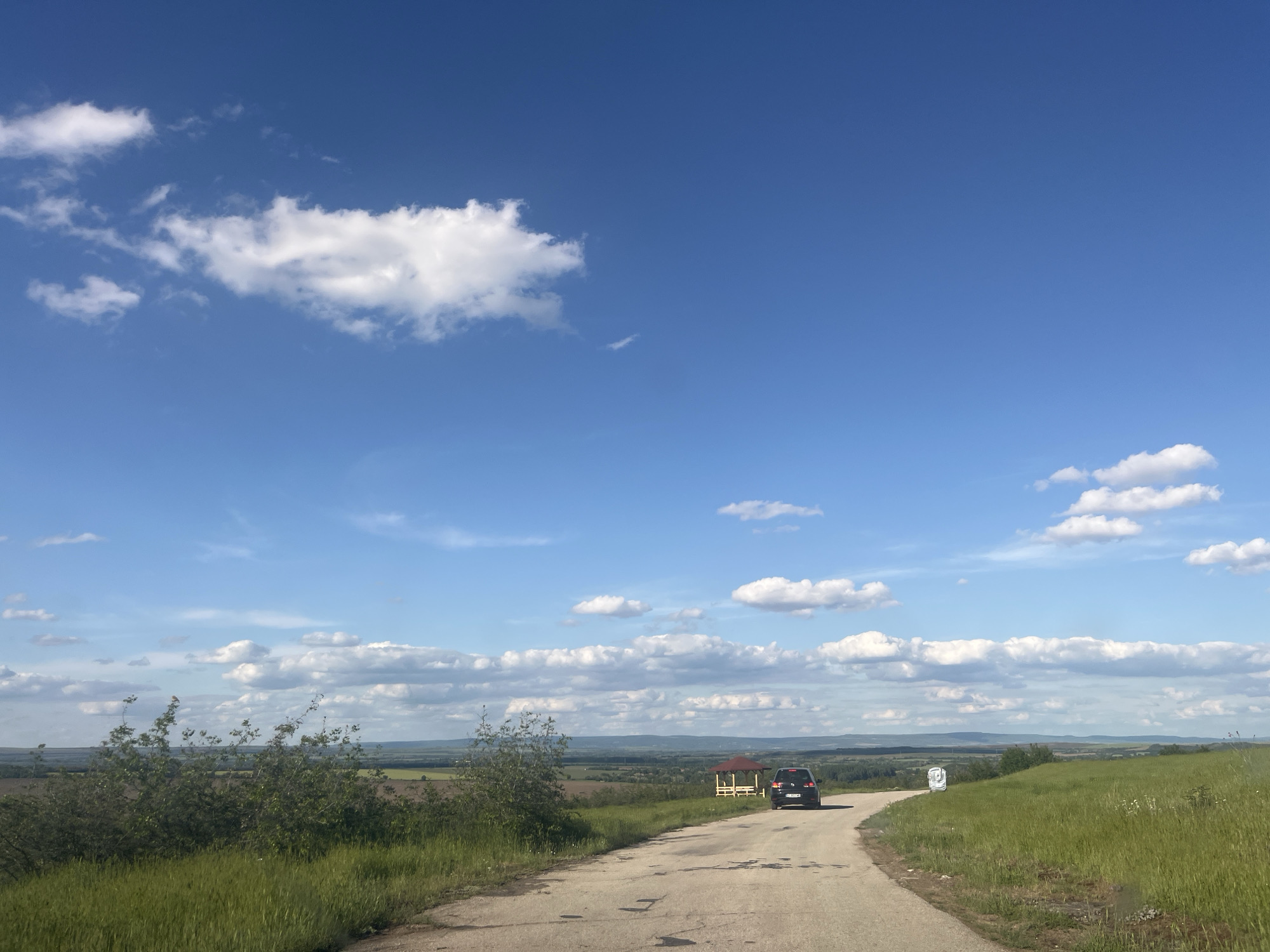 A medium shot depicts a rural scene on a sunny day in an unidentified location. The sky is a gradient of blue, with scattered cumulus clouds. A weathered asphalt road stretches into the distance, lined by green grassy verges. A dark-colored car is visible on the road, approaching a gazebo. In the background, a distant view of fields and possibly a wooded area can be discerned.