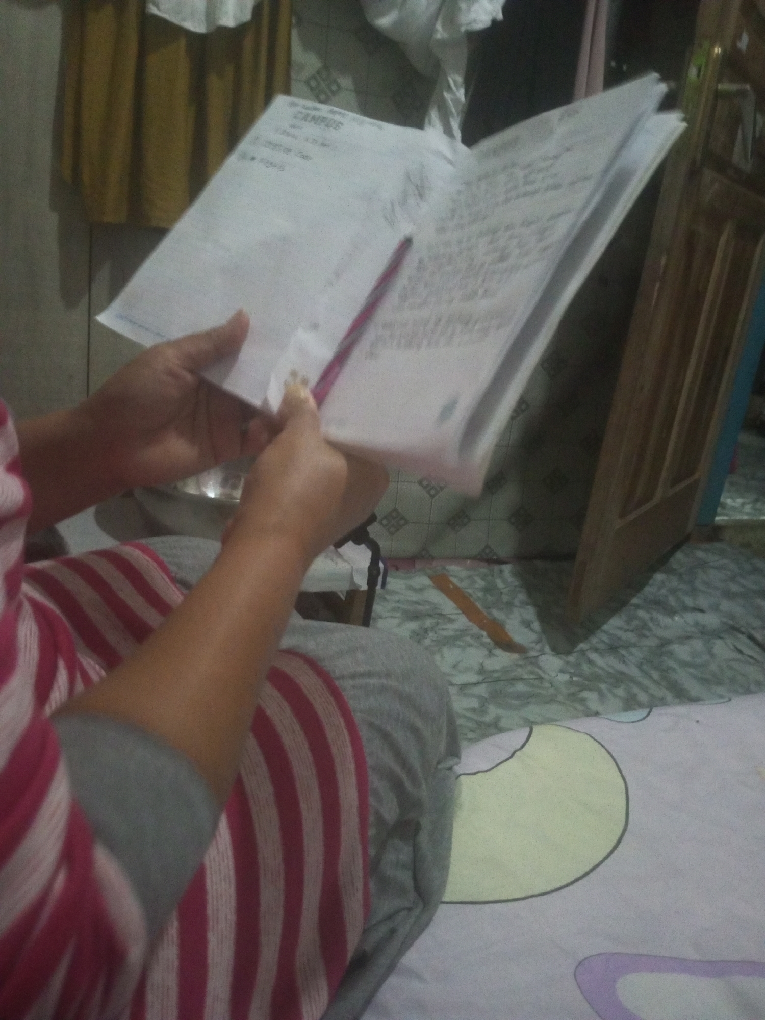 In Andongsili, Indonesia, a person's hands hold open a stapled booklet of papers. The booklet is white and appears to be filled with handwritten text. A pink pen is inserted between the pages. The person is wearing a striped shirt with pink and white stripes, and grey pants. The background shows a wall with a grid pattern, a yellow curtain, and a wooden door. The floor has a marble-like pattern.