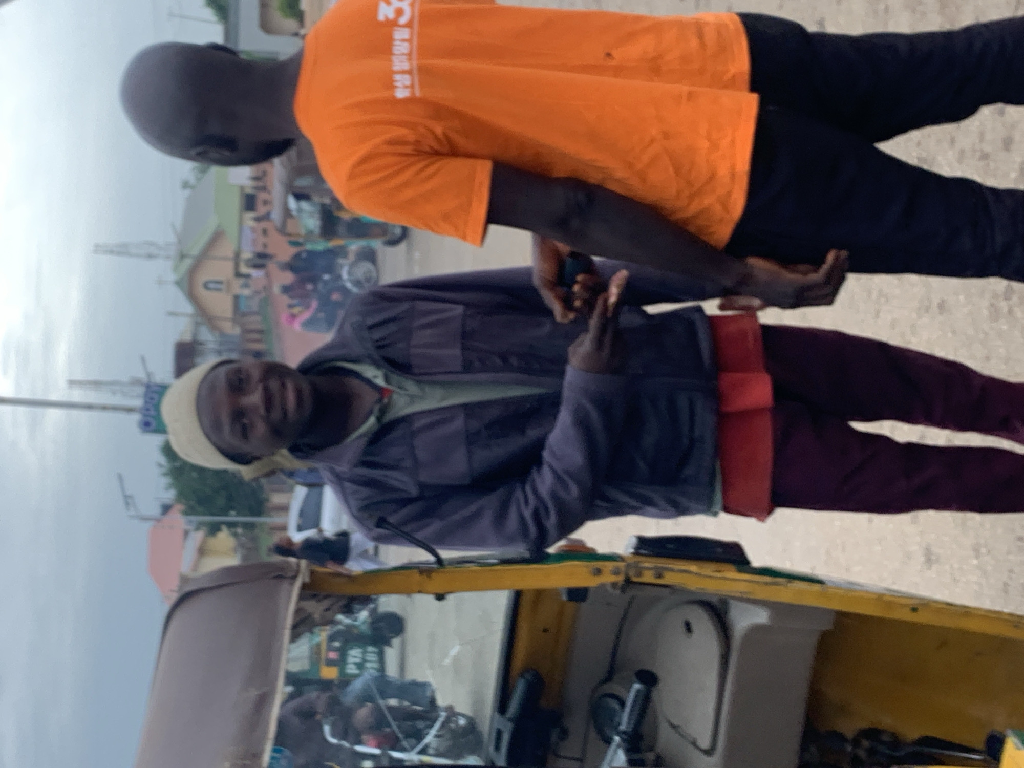 In Jos, Nigeria, two men are standing next to a yellow auto-rickshaw. The man on the left, wearing a light-colored cap and a dark jacket over a red shirt and maroon pants, is smiling and looking at the camera. He is holding a dark object in his hand, which he appears to be exchanging with the man on the right. The man on the right, facing away from the camera, is wearing an orange t-shirt with some white lettering on the back and black pants. The auto-rickshaw, positioned on the left side of the frame, has a brown canopy and visible handlebars and steering wheel. In the background, there are buildings, including a church-like structure with a green roof and a bell tower, and other vehicles, suggesting a street or marketplace setting. The sky is overcast.