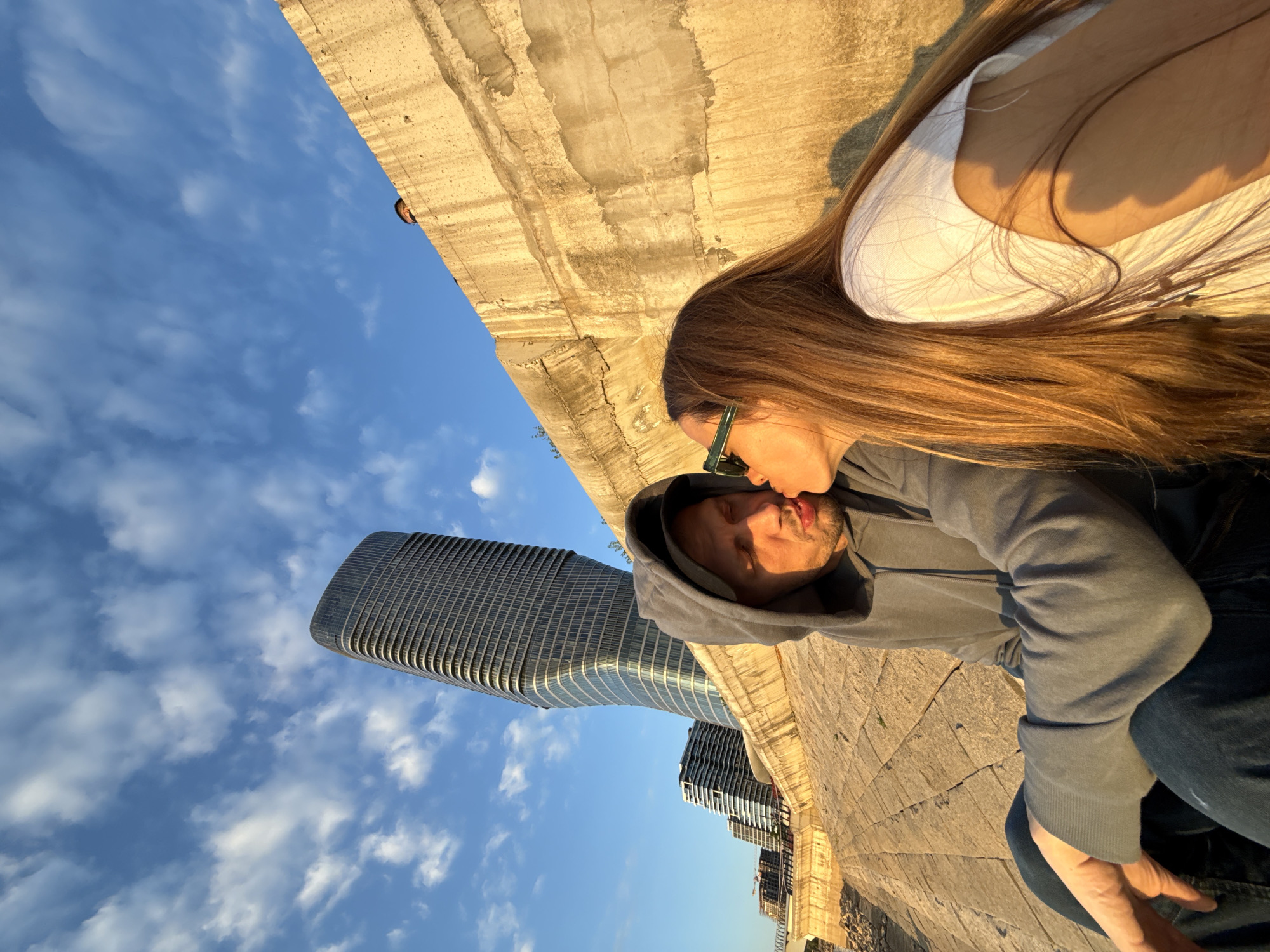 The image captures an intimate moment between a couple in an urban setting, bathed in the warm, golden light of what appears to be late afternoon or early evening. The scene is presented with an unusual high-angle perspective, making the concrete wall/ground appear on the right side and the sky and buildings on the left.
In the foreground, a woman with long, sun-kissed light brown hair is on the left, leaning in to kiss a man on the right. She wears a white top and green-framed sunglasses are visible on her head. The man, with light facial hair, has his eyes closed and lips puckered for the kiss, wearing a grey hooded sweatshirt with the hood up. A slight glimpse of his tongue is visible. Their close proximity conveys a moment of affection and romance.
In the background, a prominent modern skyscraper with a curved, reflective glass facade rises majestically into the clear blue sky, which is dotted with scattered white clouds. Other buildings are visible further in the distance. The couple is situated near a textured concrete or stone embankment or wall. A third individual is barely visible peeking over the top edge of this wall in the far upper left corner. The setting is an urban environment, identified as Belgrade, Serbia, with its distinctive architecture and clear weather. No visible text is present in the image.
