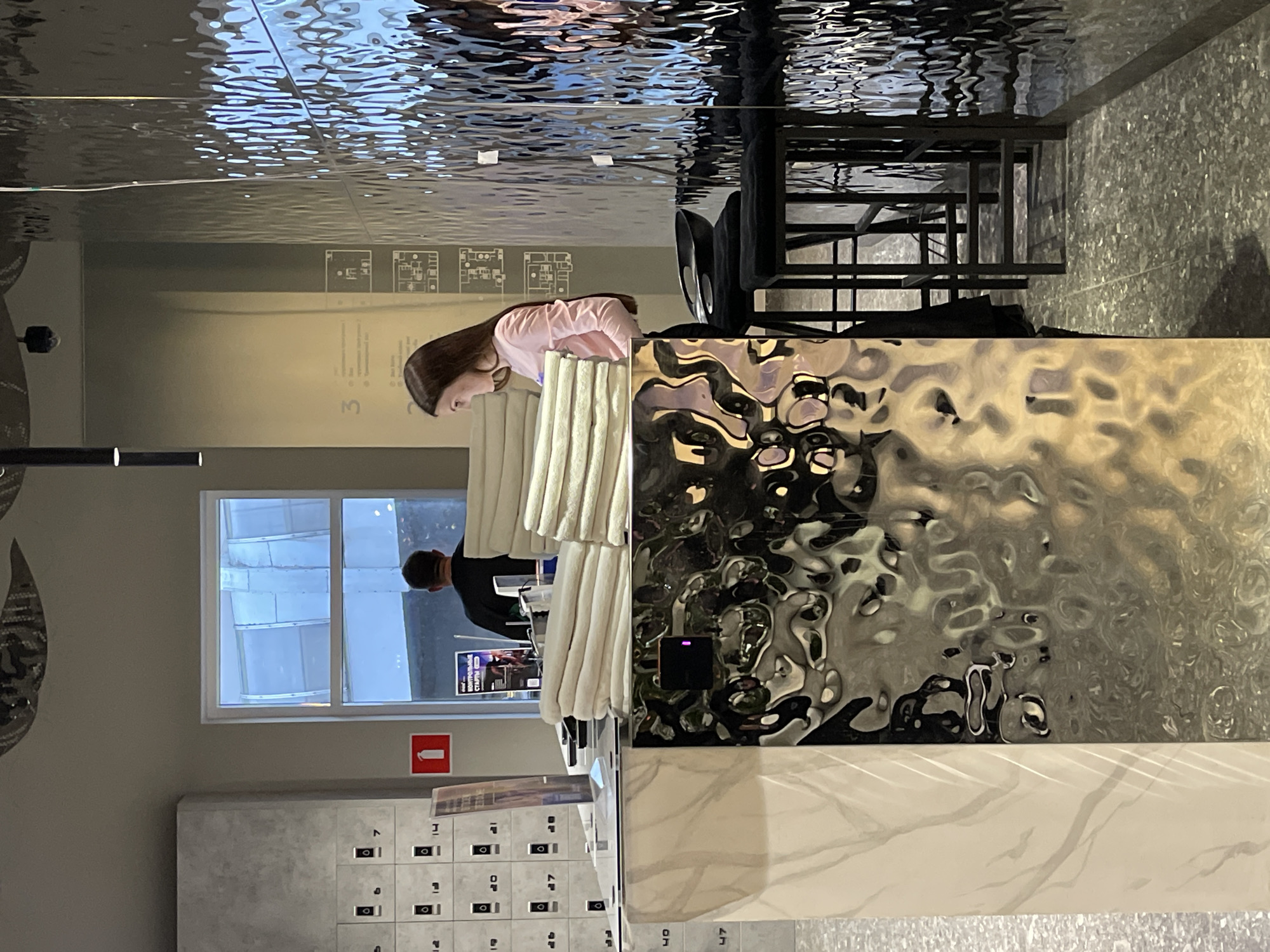The image shows the reception area of the Zaryadye Hotel in Moscow, Russia. A woman with long brown hair, wearing a pink top, is leaning over a reception desk with a textured, reflective surface. Behind the desk are white and beige cushioned panels. To the left, a window offers a glimpse of another person in a dark shirt. Below the window, there's a gray locker unit with numbered compartments and a red fire extinguisher symbol. Above, the ceiling has a reflective, textured surface. In the background, there are tables and chairs. The overall lighting is bright.