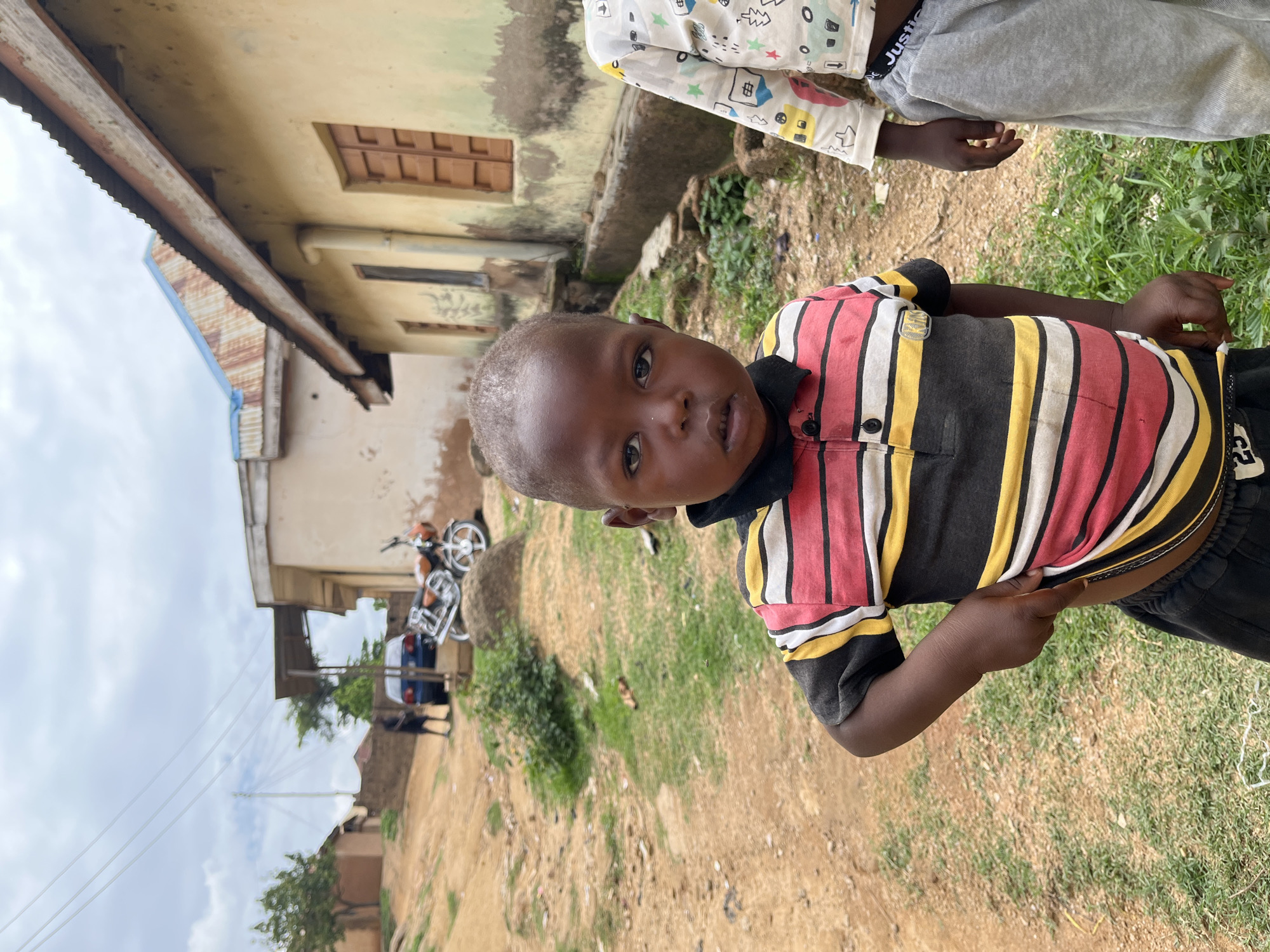 The image depicts an outdoor scene in what appears to be a residential or rural area within the city of Jos, Nigeria, under an overcast sky.
In the foreground, a young Black male child, approximately toddler age, stands facing the camera. He has a very short dark haircut and wears a black-collared polo shirt with horizontal stripes in red, white, yellow, and black, along with black shorts. His left hand is tucked into the waistband of his shorts, slightly pulling down the front of his shirt. His eyes are open and directed at the viewer. To his right, the partial figure of another child is visible, wearing a white long-sleeved shirt with multi-colored patterns and grey sweatpants.
The background features an unpaved, reddish-brown dirt path or ground with patches of green grass and sparse vegetation. On the right side, a single-story building with light yellow walls, a corrugated metal roof, and several brown wooden shuttered windows is visible. A drainpipe runs vertically along its wall. Further back on the left, another building with a reddish roof is partially visible, and beyond that, another light-colored structure with a blue/rusted corrugated roof.
In the mid-ground, a dark blue sedan is parked on the dirt, with two blurry figures standing nearby. To the right of the car, a silver and orange/brown motorcycle is parked on a raised dirt mound. Multiple thin utility wires traverse the upper portion of the frame against the light grey, cloudy sky.