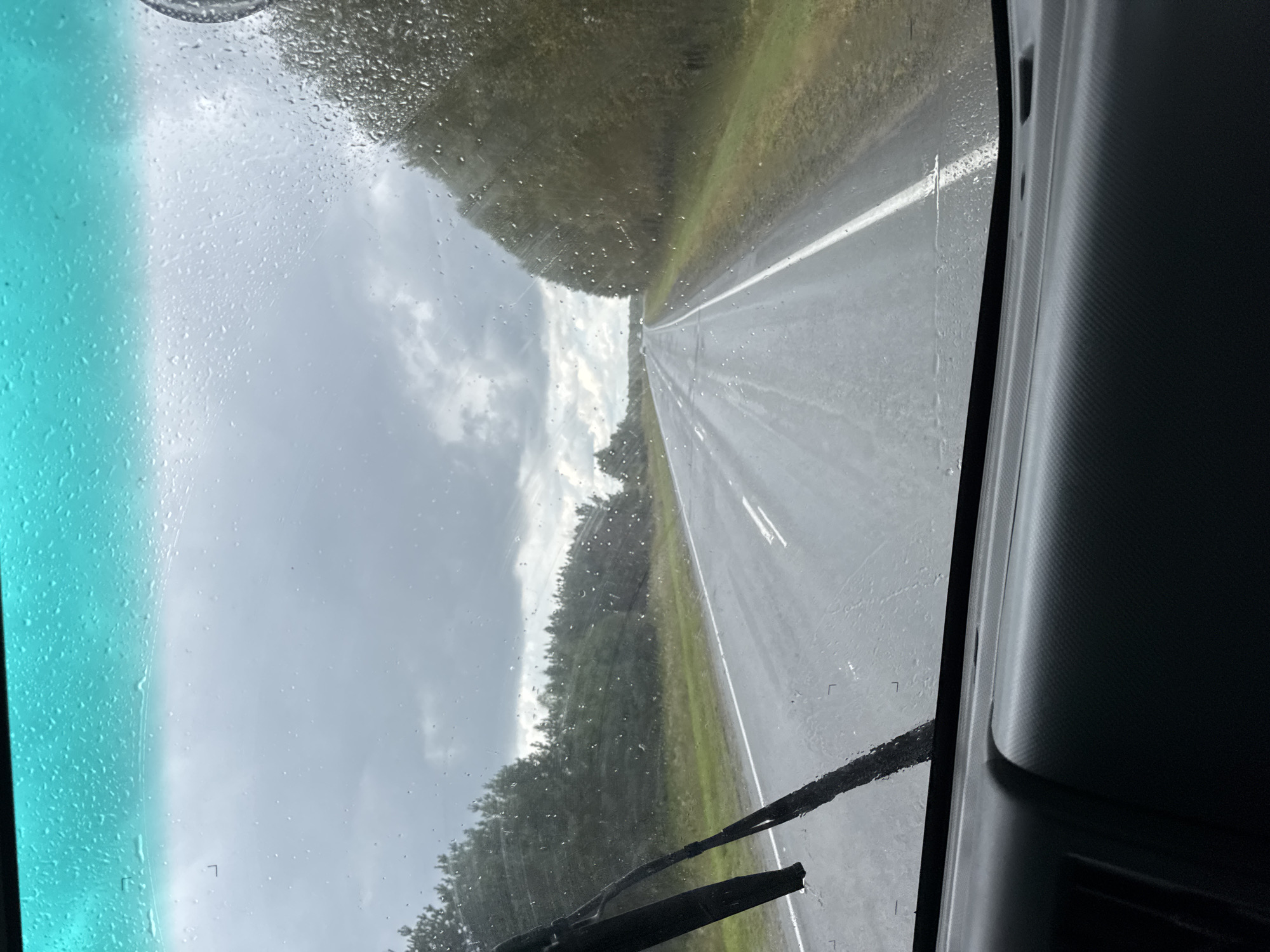 An exterior, eye-level, medium shot shows a view through a car windshield on a rainy day. The windshield is covered with raindrops and streaks, obscuring the view slightly. In the foreground, the black windshield wiper is visible on the left side. The road ahead is wet and appears to be a highway with white lines marking the lanes. Trees line both sides of the road, and the sky is overcast with dark clouds. The interior of the car's dashboard is visible at the bottom of the frame, showing a textured black surface. The overall scene conveys a sense of driving in inclement weather.