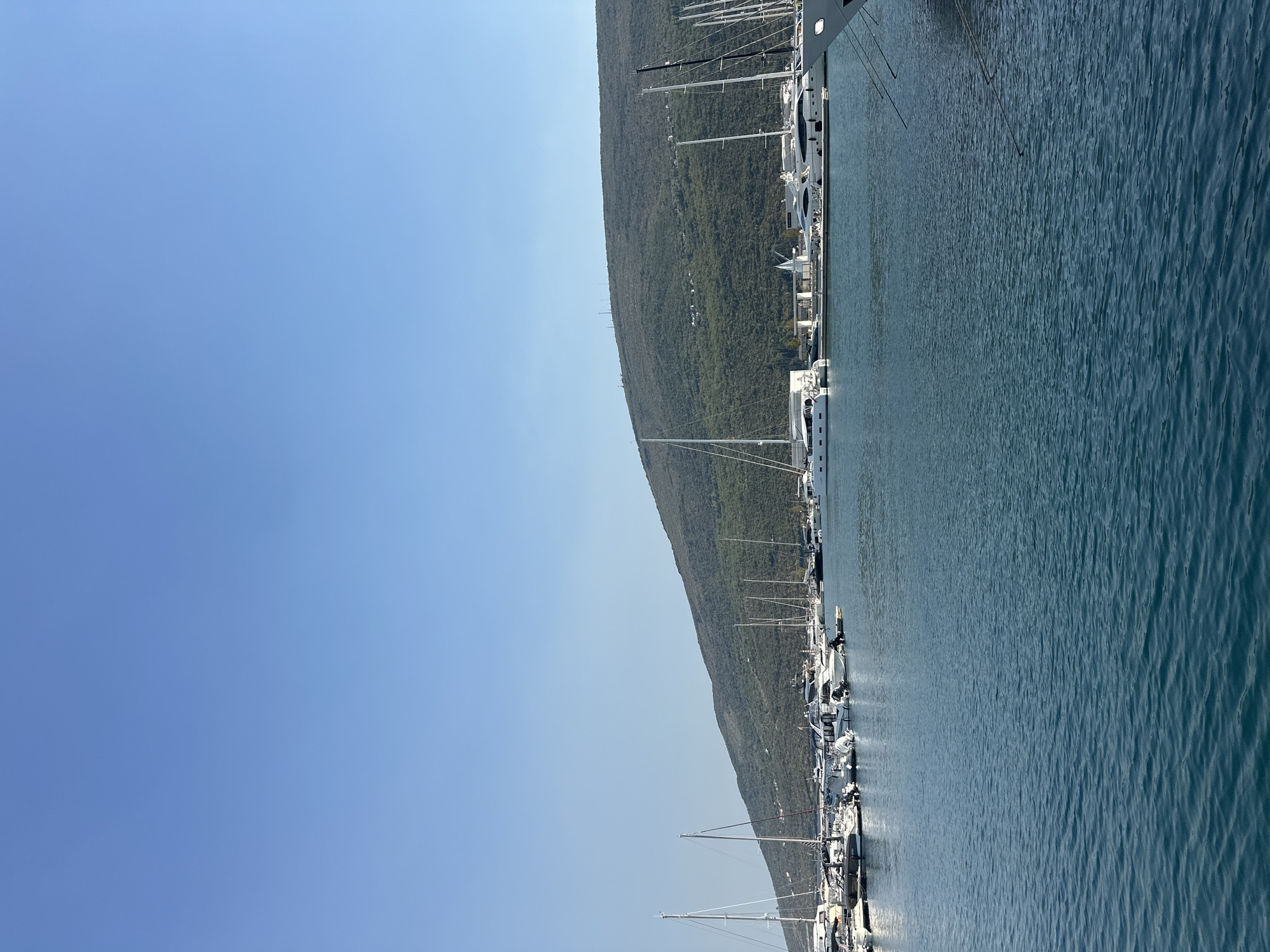 The image shows a bright, sunny day at a marina in Kumbor, Montenegro. The sky is clear and blue, with a gentle haze near the horizon. The foreground is dominated by the calm, deep blue water of the marina, with small ripples on the surface.
A fleet of various boats, including sailboats and yachts, are docked in the marina. The boats are white and appear to be well-maintained, with tall masts of the sailboats reaching towards the sky. Some boats have dark hulls or accents, adding contrast to the scene. The marina is lined with docks and walkways.
In the background, a large, lush green mountain slopes down towards the water, its surface covered in dense vegetation. Small structures and possibly some scattered houses are visible on the mountainside. The overall impression is one of a tranquil and picturesque coastal location, characteristic of the Bay of Kotor. There are no people clearly visible in the image.