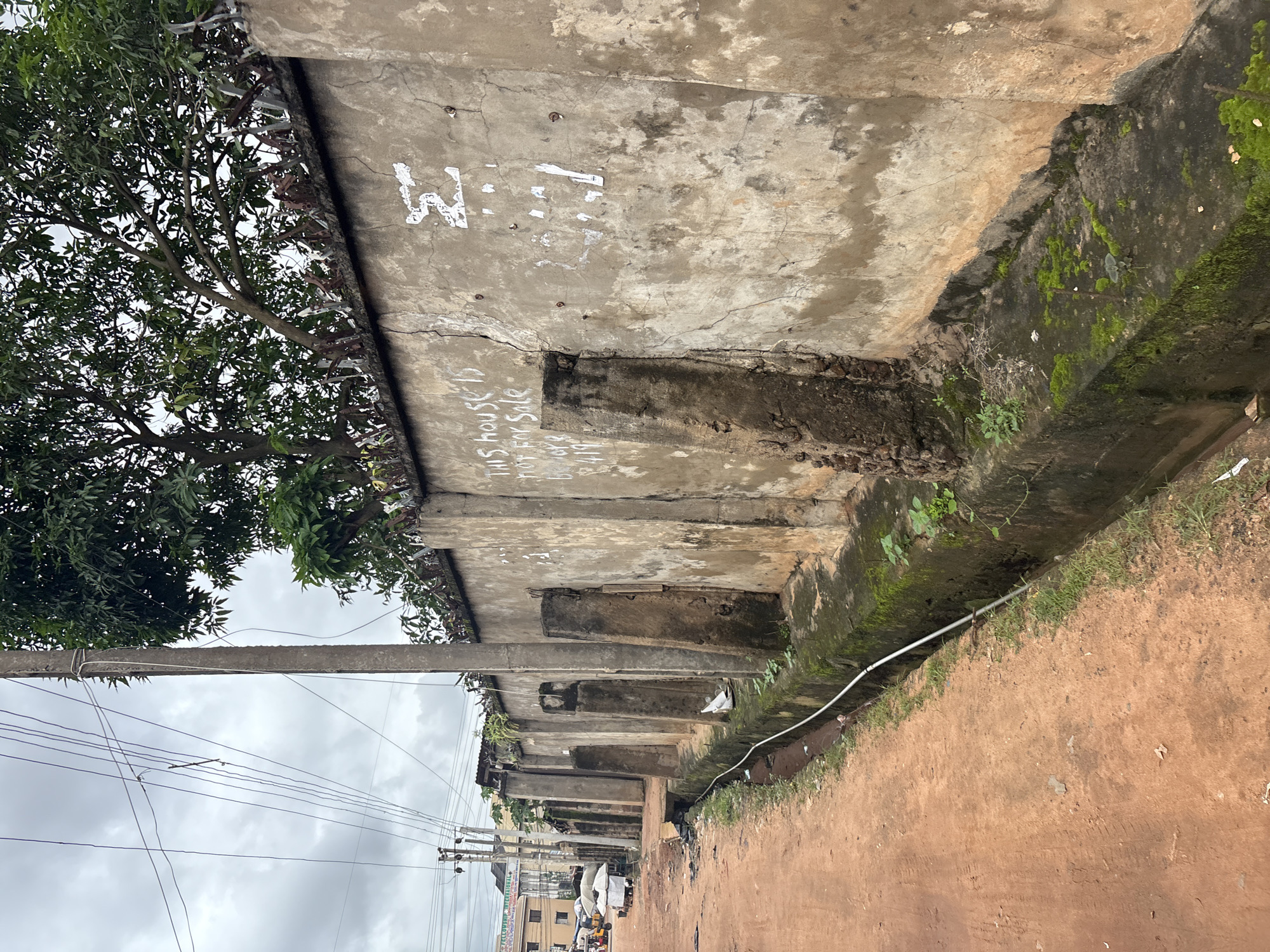This image depicts a street scene in Nkpor, Nigeria. A rough, reddish-brown dirt road runs along the left side of the frame, with a concrete drainage ditch bordering it. On the right, a weathered concrete wall runs the length of the scene, showing signs of age and damage, with visible cracks and patches of discoloration. The wall is topped with barbed wire for security. Graffiti, including "THIS HOUSE IS NOT FOR SALE", is visible on the wall. A concrete utility pole with numerous overhead wires stands prominently in the left-center of the image. Behind the wall, trees with lush green foliage can be seen. In the background, a few buildings and market stalls with umbrellas suggest a commercial or residential area. The sky is overcast, indicating a cloudy day. No people are clearly visible in the immediate foreground or performing any actions.