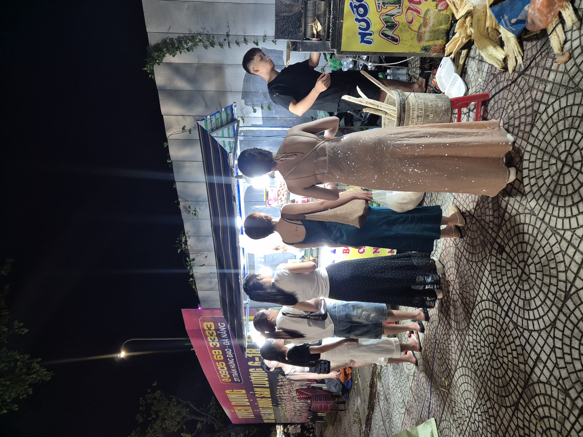 In the city of Đà Nẵng, Vietnam, at night, several people are gathered at a street-side vendor. The vendor appears to be selling sugarcane juice. A young man in a black t-shirt and shorts operates a sugarcane juicing machine. Five women are in line, waiting to be served. One woman, wearing a sequined dress, is closer to the juicing machine, while another woman in a teal dress stands beside her. The women are dressed in casual attire, with one wearing a long black skirt with white polka dots and another in denim shorts. The ground is paved with a circular mosaic pattern. A large pink banner advertising "TUYÊN DỤNG 0905 69 3333" and an address in Đà Nẵng is visible in the background. The scene is illuminated by overhead lights and the glow from the vendor's stall. Sugarcane stalks are piled next to the juicing machine, and there are cups and other supplies. The overall impression is that of a busy nighttime street food scene.