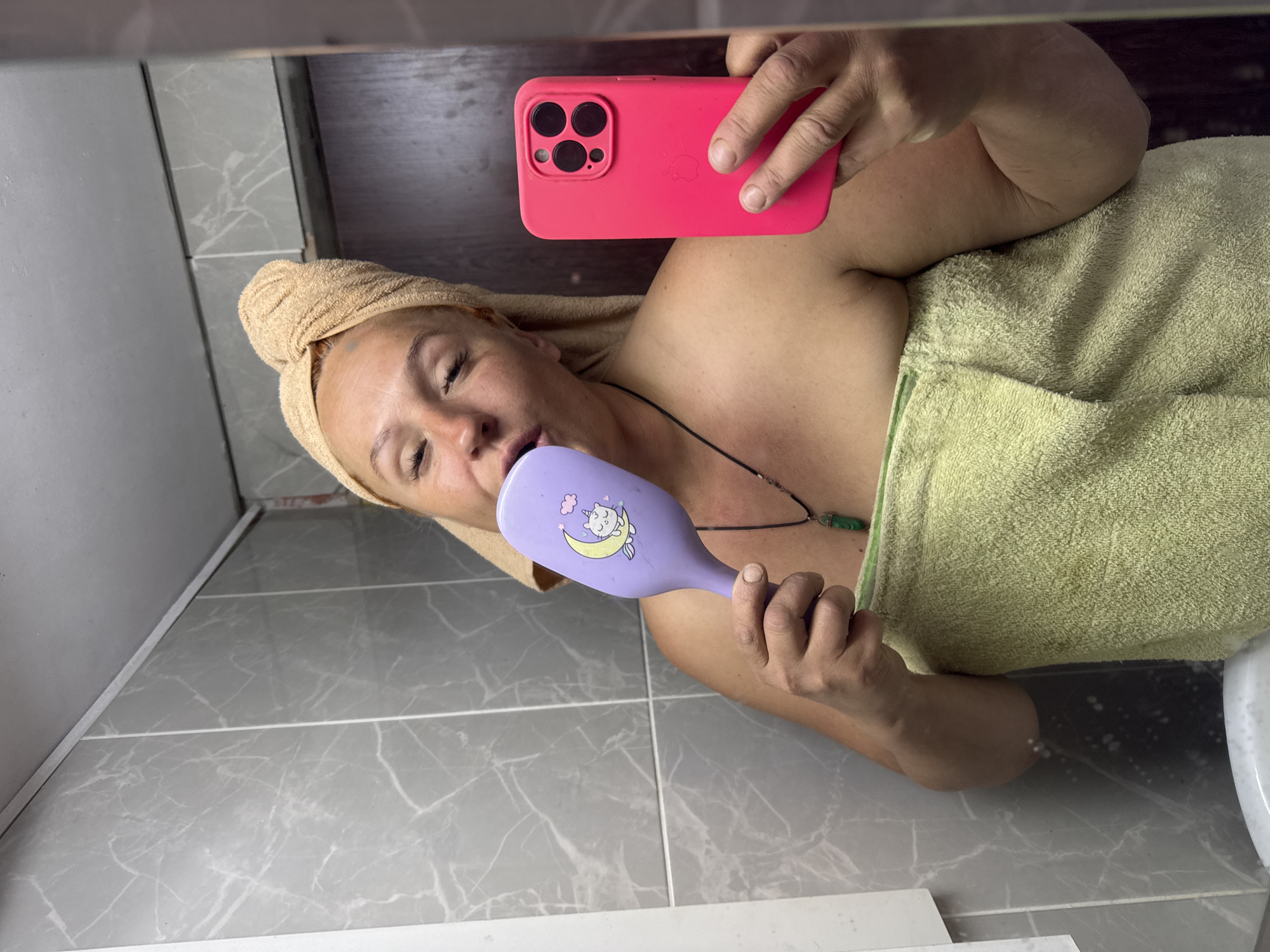 A woman, wrapped in towels, is taking a selfie in a bathroom. She has a light-colored towel wrapped around her head and a lighter green towel wrapped around her body. She is holding a purple hairbrush with a cartoon cat on it to her mouth and a bright pink smartphone in her right hand, which she is using to take the photo. The bathroom appears to have gray, marble-patterned tiles on the wall. The lighting suggests it is daytime. The location is Chaikovsky, Russia.
