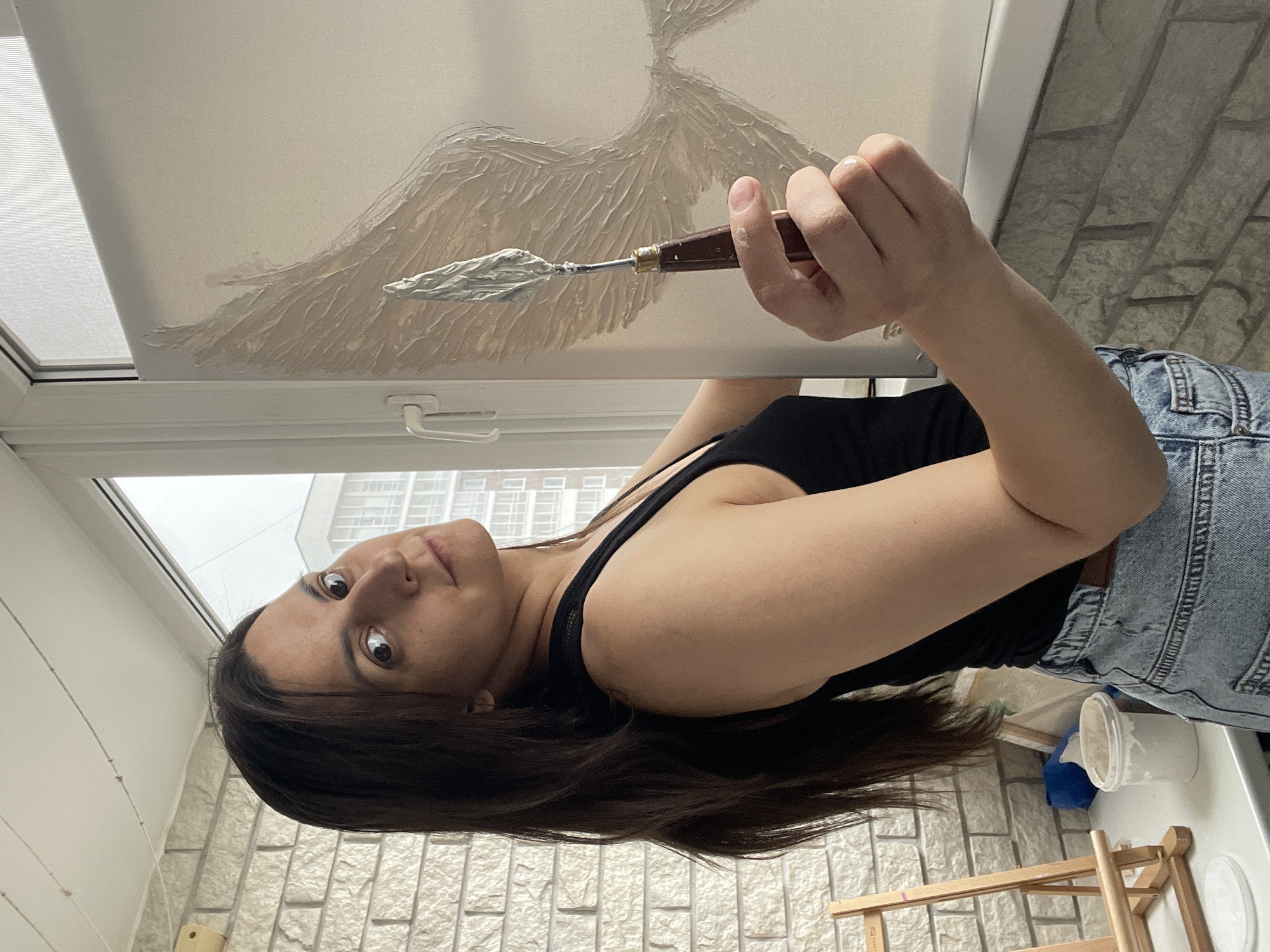 A woman in a black tank top and light blue jeans is painting on a white canvas near a window. She is holding a palette knife with light-colored paint on it. The painting appears to be an abstract design with textured paint strokes. The location seems to be indoors, possibly an apartment, with a white brick-patterned wall. There's a small wooden easel and a white container nearby. The city and region are unknown.