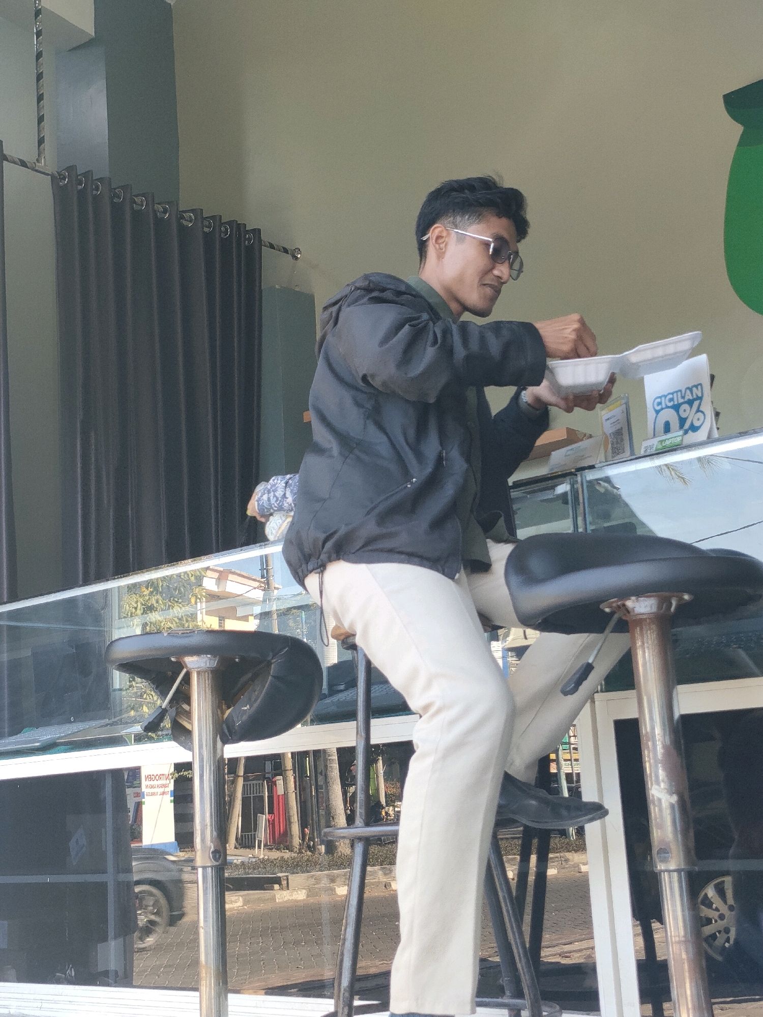 A man sits on a stool at a counter in Yosowilangun, Indonesia, holding a white takeout container with food in his right hand and a utensil in his left, appearing to be eating. He wears a dark jacket over a green shirt and light-colored pants. Sunglasses are on his face. Behind him, dark curtains are visible. The counter has a glass surface reflecting the street outside, showing a road with parked cars and buildings. A sign with "CICILAN 0%" and "LAPTOP" is also visible on the counter. Two chrome-legged stools with black seats are positioned at the counter. The environment suggests an outdoor dining or retail setting with a clear view of the street.