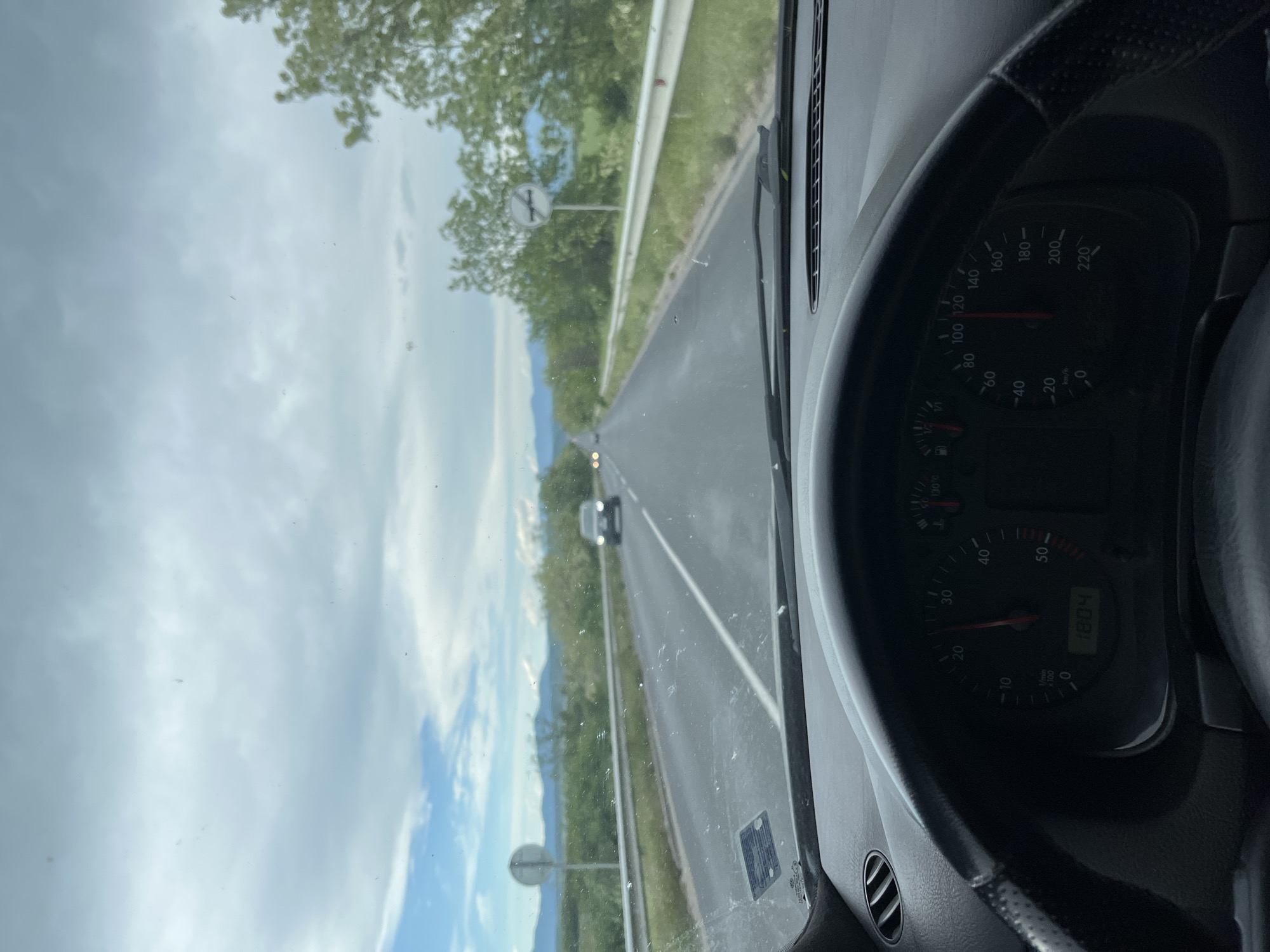 A view from inside a car driving on a road outside of Dolna Mahala, Bulgaria. The car's dashboard and speedometer are visible on the right, with the speedometer indicating a speed of approximately 120 km/h. The road stretches ahead, lined with trees and fields on either side. Another car is visible in the distance. The sky is overcast with clouds. A sign indicates a junction ahead.