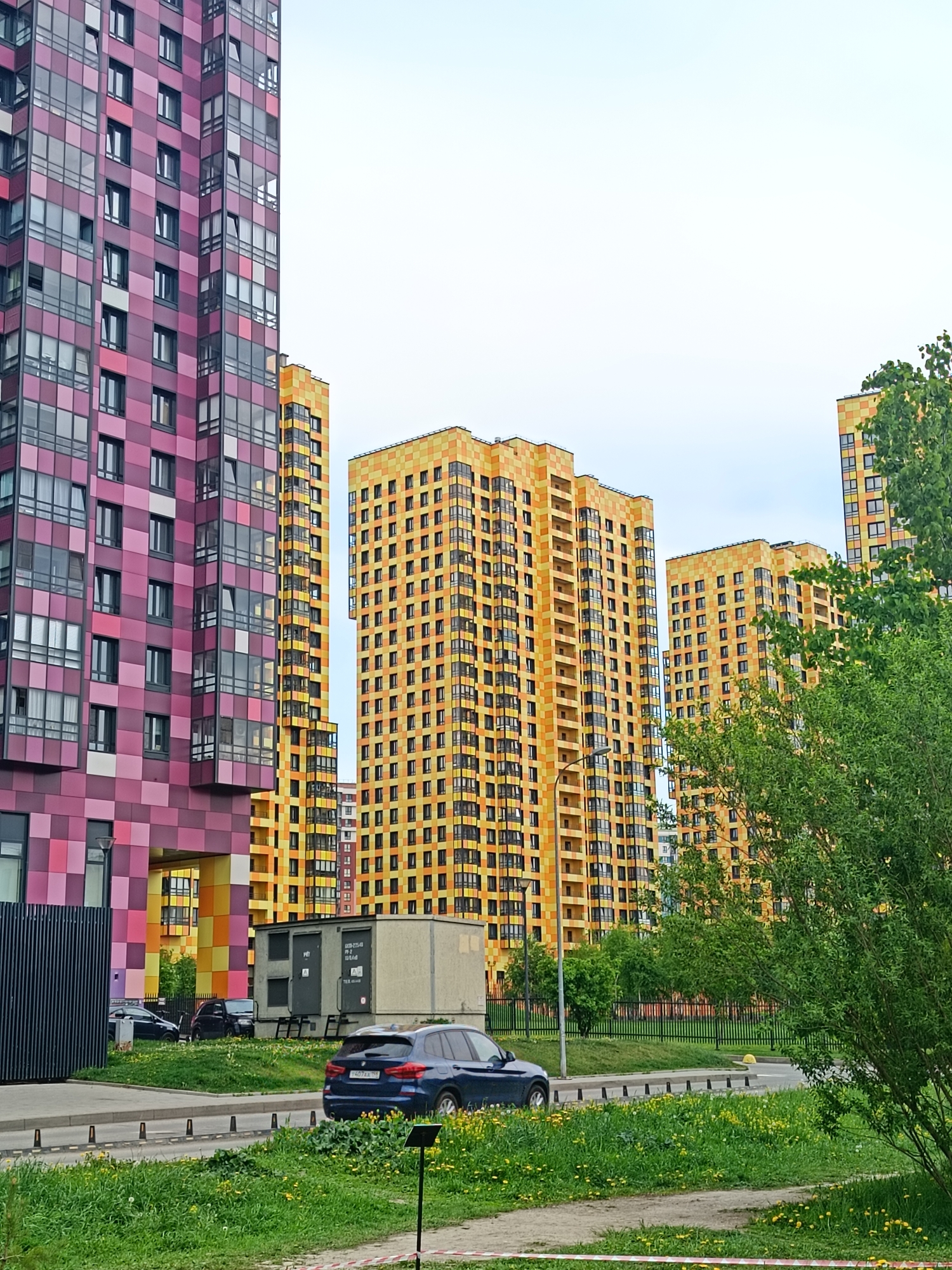 The image shows modern apartment buildings in Saint Petersburg, Russia. A tall building with a pink and purple checkered facade stands on the left side of the frame. To the right, three yellow and orange checkered apartment buildings rise into the sky. In the foreground, there's a green lawn with yellow flowers. A dark blue SUV is parked on the side of the road. Behind the SUV are several containers and other parked cars. A green tree is visible on the right side of the frame. The sky is overcast.