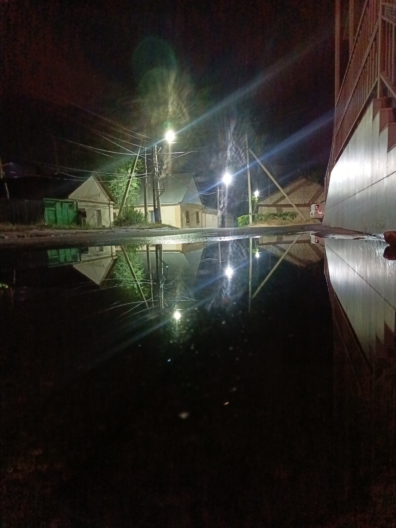 This media file captures a nighttime street scene in Shymkent, Kazakhstan, with a focus on a large puddle reflecting the surroundings. The scene is illuminated by streetlights, creating bright reflections in the water.
The subjects include a row of simple, light-colored buildings with gabled roofs, and a bright green dumpster. Utility poles with numerous wires are visible, suggesting an urban or semi-urban environment. To the right, a portion of a building with a light-colored wall and a metal railing, possibly a staircase or balcony, is present. The puddle dominates the foreground, serving as a mirror to the scene above, distorting the shapes and colors of the buildings and lights.
The time of day is clearly night, as indicated by the darkness of the sky and the artificial illumination. The presence of a large puddle suggests recent rainfall or damp conditions, and the overall atmosphere is one of quietude and perhaps a touch of melancholy associated with a wet, dark night. There is no visible text in the image. The location is identified as Shymkent, Kazakhstan, but specific landmarks are not apparent beyond the general context of a residential or commercial street.