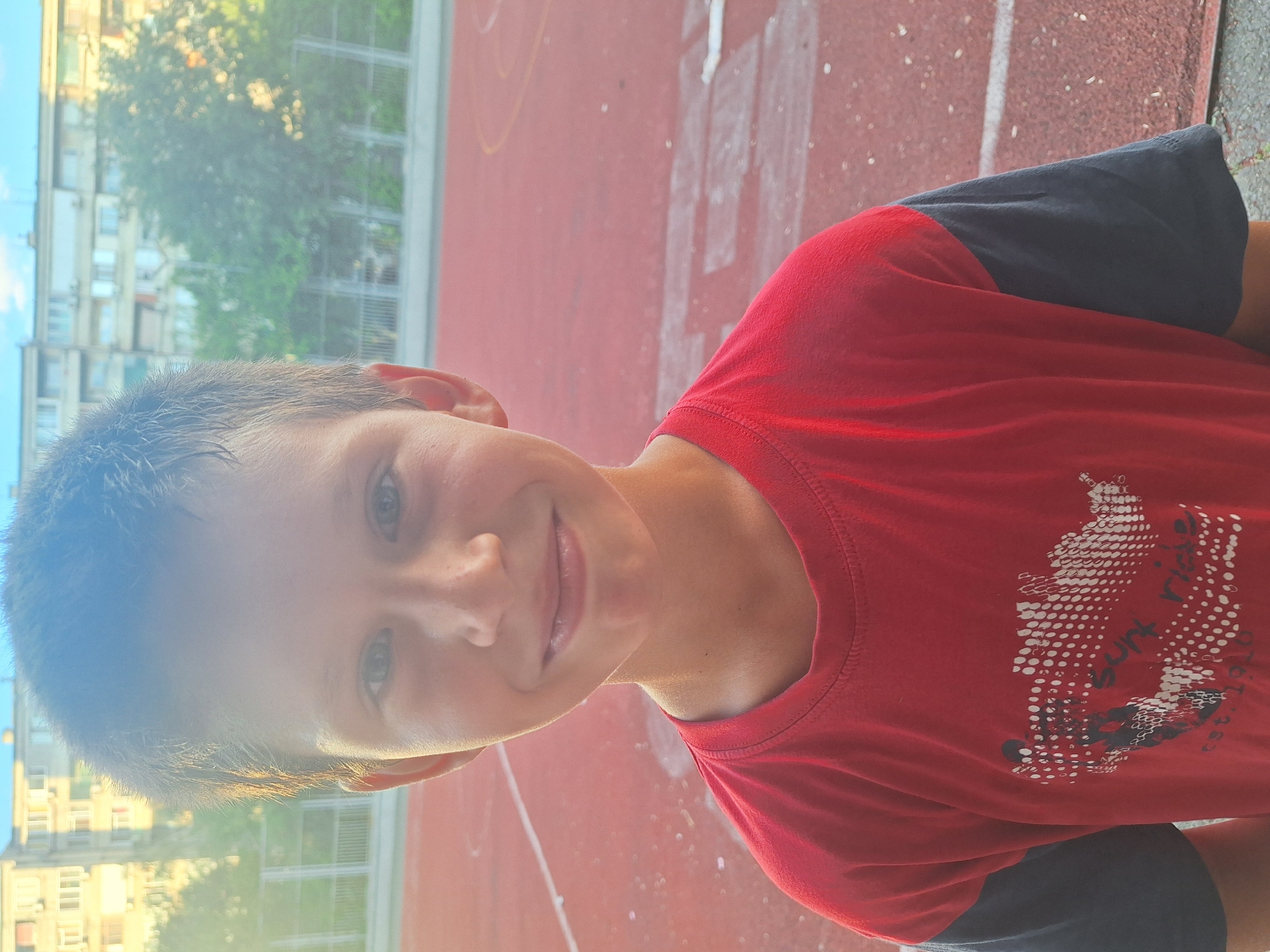A young boy with short brown hair and a light complexion smiles at the camera. He is wearing a red t-shirt with grey sleeves and a graphic print. The boy is standing on a red athletic track with white lines. In the background, there are trees and a light-colored apartment building. The lighting suggests it is daytime, possibly late afternoon due to the warm tones and lens flare. The scene appears to be an outdoor recreational area, common in urban settings like Yambol, Bulgaria.