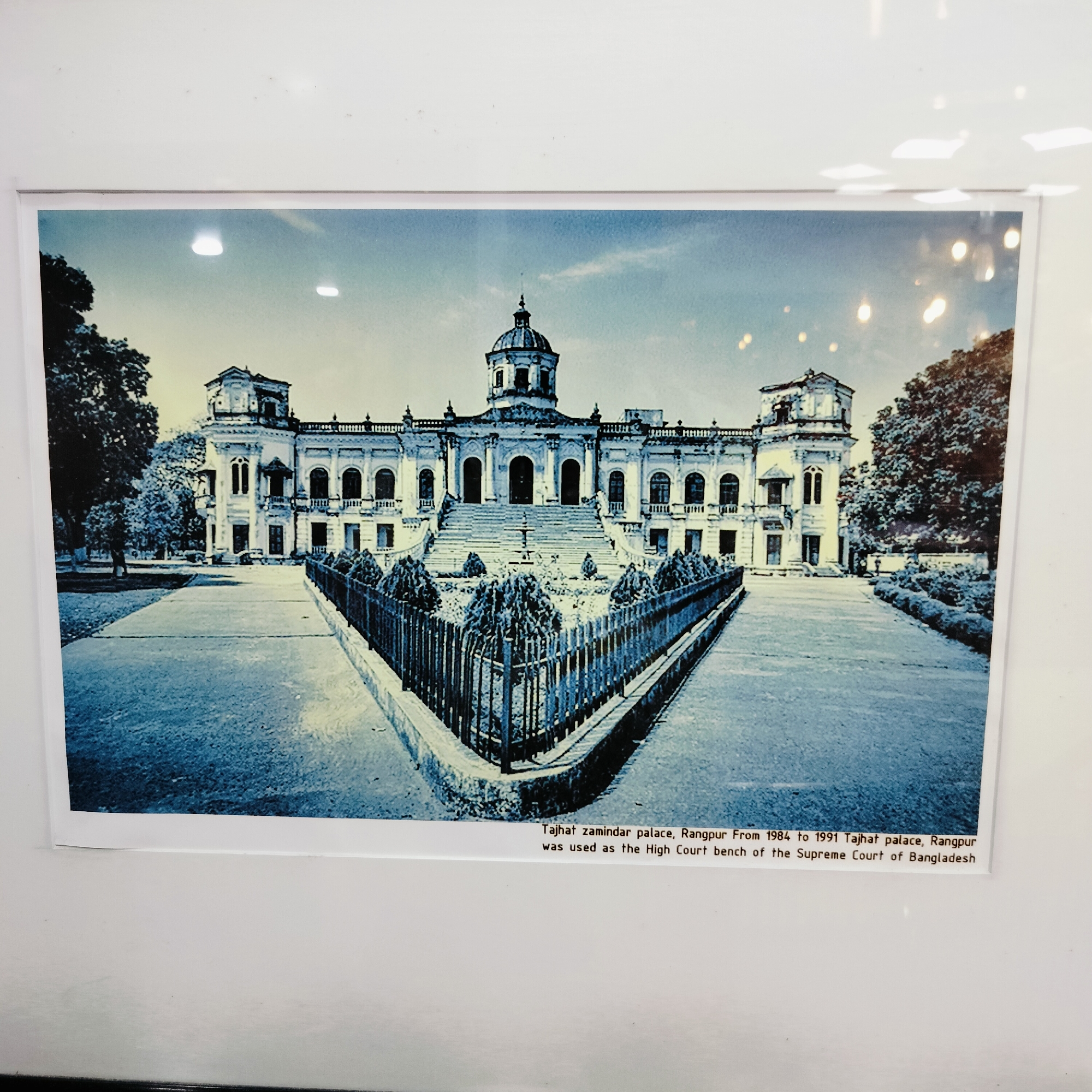 The image displays a physical print of the Tajhat Zamindar Palace in Rangpur, Bangladesh. The photograph itself exhibits a strong blueish, almost monochromatic, tone, lending it a historic or stylized aesthetic. The central subject is the grand, symmetrical palace, characterized by a prominent central dome, flanked by two side towers, numerous arched windows, balconies, and intricate architectural embellishments. A broad, multi-tiered staircase leads up to the main entrance of the building.
In the foreground, a paved pathway guides the eye towards the palace. This pathway is bordered by a V-shaped garden plot, meticulously arranged with small, manicured bushes, and enclosed by a dark metal fence. Large, leafy trees frame both sides of the palace in the background. The sky above is bright with scattered clouds, indicating that the scene was captured during daytime. Notably, several reflections from overhead light sources are visible on the surface of the print, suggesting it is displayed behind glass or a protective covering.
No people or discernible activities are present in the image, allowing the focus to remain on the architectural grandeur of the palace.
The text visible beneath the image provides contextual information: "Tajhat zamindar palace, Rangpur From 1984 to 1991 Tajhat palace, Rangpur was used as the High Court bench of the Supreme Court of Bangladesh". Although the provided location context for the media file is Bhola, Bangladesh, the subject of the photograph is specifically identified as being located in Rangpur.