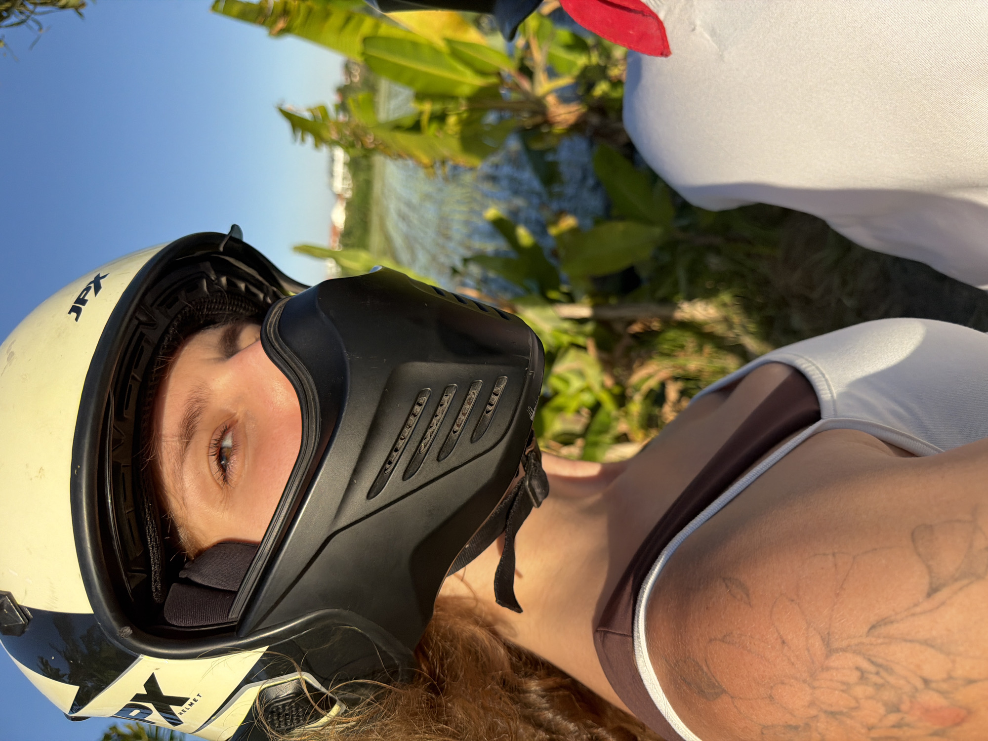 A person wearing a motorcycle helmet is visible in the foreground. The helmet is white with black accents and a black face mask. Only one eye of the person is visible inside the helmet. The person is wearing a two-tone swimsuit with brown and white colors. On the shoulder area, a tattoo is visible.
In the background, there is a body of water and green vegetation. The sky is light blue.
While not explicitly stated, the prompt references North Kuta, Indonesia. The scene suggests a tropical or subtropical environment. The presence of a motorcycle helmet implies the subject is likely riding or preparing to ride a motorcycle.