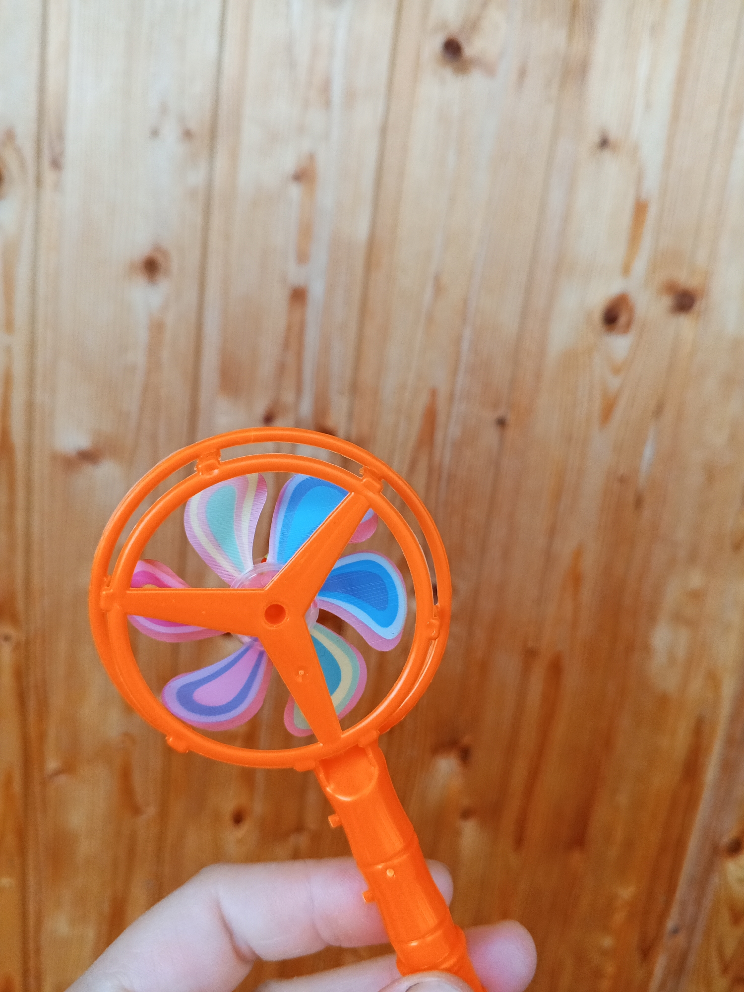 An orange plastic pinwheel toy with colorful blades is held in a person's hand against a backdrop of wooden planks. The wooden planks are oriented vertically and have a warm, brown tone with visible knots and grain. The pinwheel's circular frame is bright orange, and its four blades are patterned with stripes of pink, light blue, and yellow. The toy is positioned in the lower left portion of the frame, with the hand and handle extending downwards. The image is taken from a slightly elevated perspective.