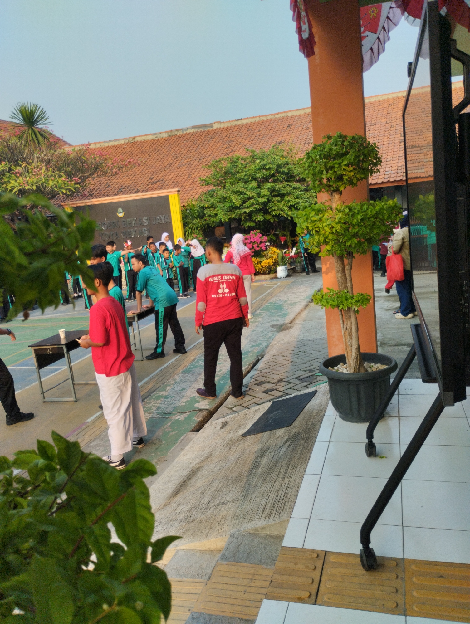 The image depicts an outdoor scene at what appears to be a school in the city of Bekasi, Indonesia. In the background, a building with a red tiled roof and a yellow facade with text "NEGERI BEKASI JAYA KOTA BEKASI" is visible. Several young people, most wearing teal uniforms with black pants or skirts, are gathered in what looks like an open area or courtyard, possibly for an event or activity. Some are standing in groups, while others appear to be participating in a game or demonstration.
In the foreground, a person in a red shirt and white pants stands near a table with a cup on it. Another person in a red shirt with white sleeves and text that appears to read "GUGUS DEPAN" is also visible. A potted plant with green leaves is placed near a large black monitor stand on wheels. The ground is a mix of concrete paving stones and textured yellow tactile paving. The overall atmosphere suggests a lively daytime gathering, likely a school event in Bekasi.