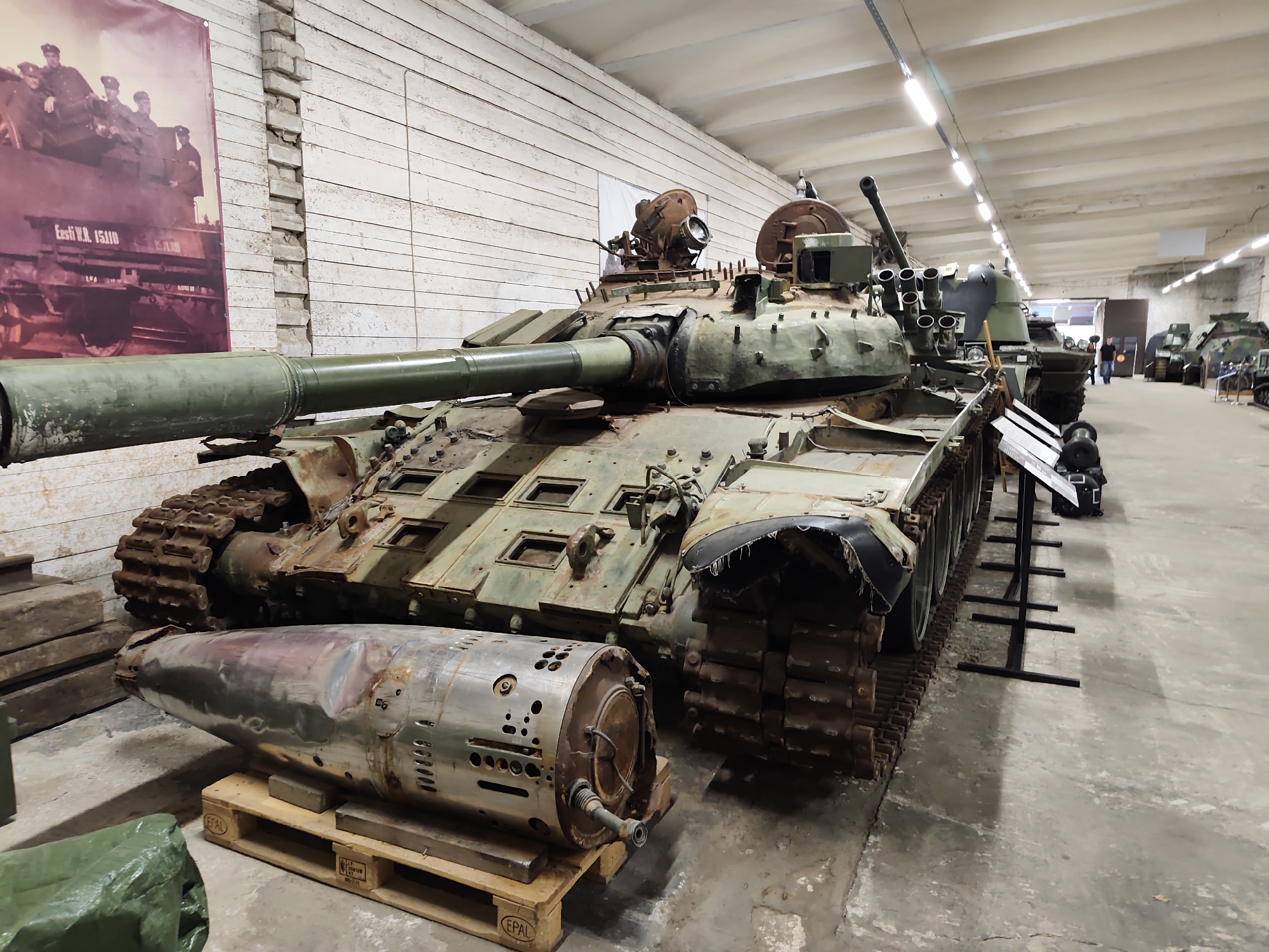 The image depicts a museum interior with a prominent tank as the central subject. The tank, appearing weathered and aged, is positioned indoors in what seems to be a large hangar or exhibition hall. The setting is industrial, with a concrete floor and a white, slatted ceiling. In the background, other military vehicles, including tanks and possibly an armored personnel carrier, are visible, suggesting a military museum or a collection of historical equipment.
In the foreground, a large, metallic object, likely a projectile or a component of artillery, rests on a wooden pallet. This object is cylindrical with a pointed end and shows signs of wear and tear. The tank itself is a complex machine with tracks, a large gun barrel, and various mounted components, including what appear to be multiple smaller cannons or rocket launchers on its turret. The overall condition of the tank suggests it's a display piece rather than an operational vehicle.
On the wall behind the tank, a large photograph or banner is displayed. It shows a group of people in uniform on top of a military vehicle. Text visible on the banner reads "Eesti V.R. 15110".
The lighting in the image is artificial, coming from overhead fixtures. The scene conveys a sense of history and preservation, with the exhibits likely representing past conflicts or military capabilities. The focus is on the artifacts themselves, with no active event or human interaction clearly depicted, other than the distant figures of a few visitors in the background. The overall impression is one of a static display in a controlled environment.
