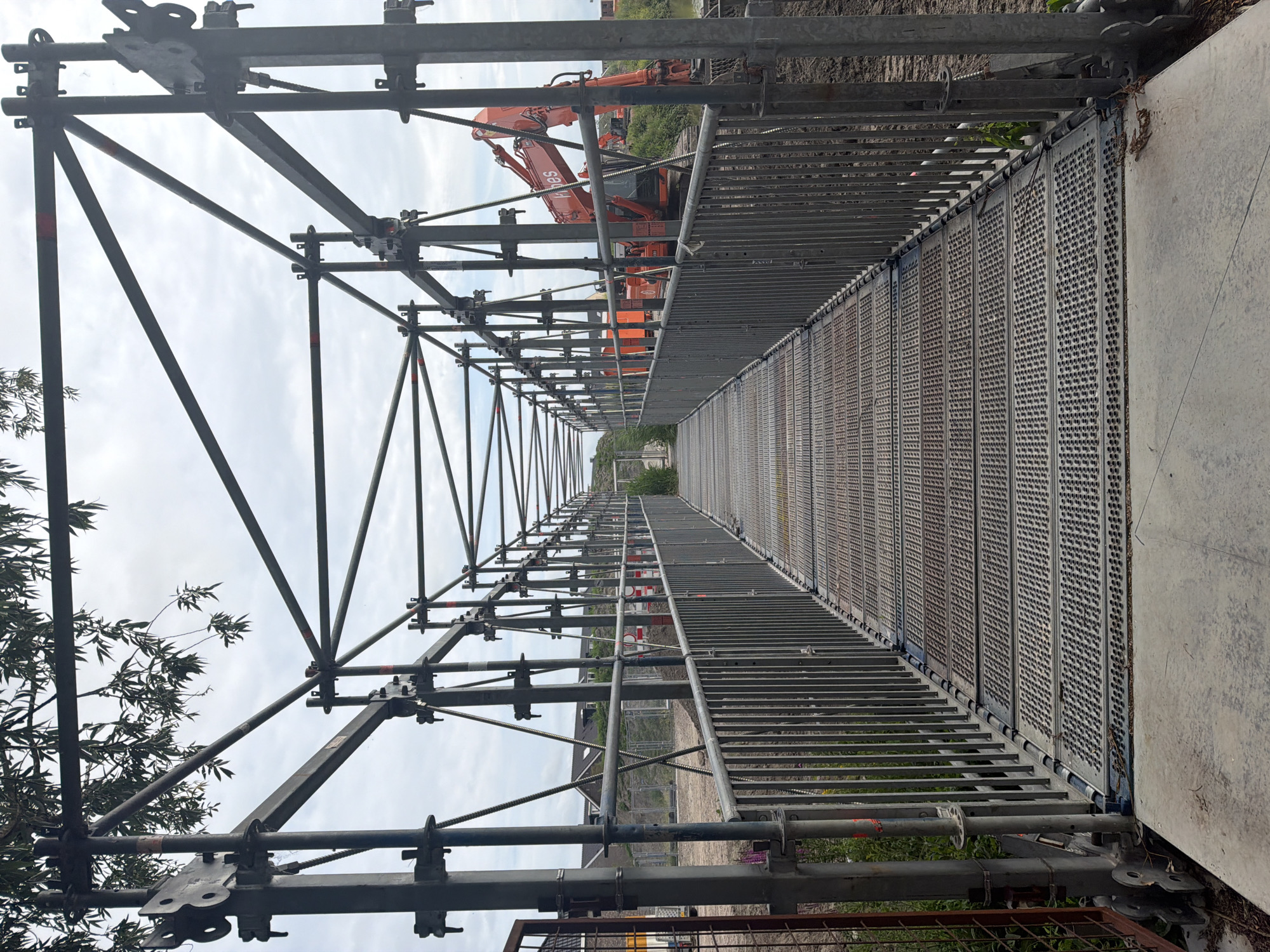 This is a photograph taken in Rijswijk, Netherlands, showing a temporary pedestrian walkway constructed with scaffolding. The walkway is made of metal grating and is bordered by metal railings. The scaffolding structure extends above the walkway, forming a tunnel-like effect. In the background, an orange excavator is visible, suggesting ongoing construction or renovation work. The sky is overcast with clouds. The overall scene depicts a utilitarian temporary structure in an outdoor, possibly industrial or construction site, environment.