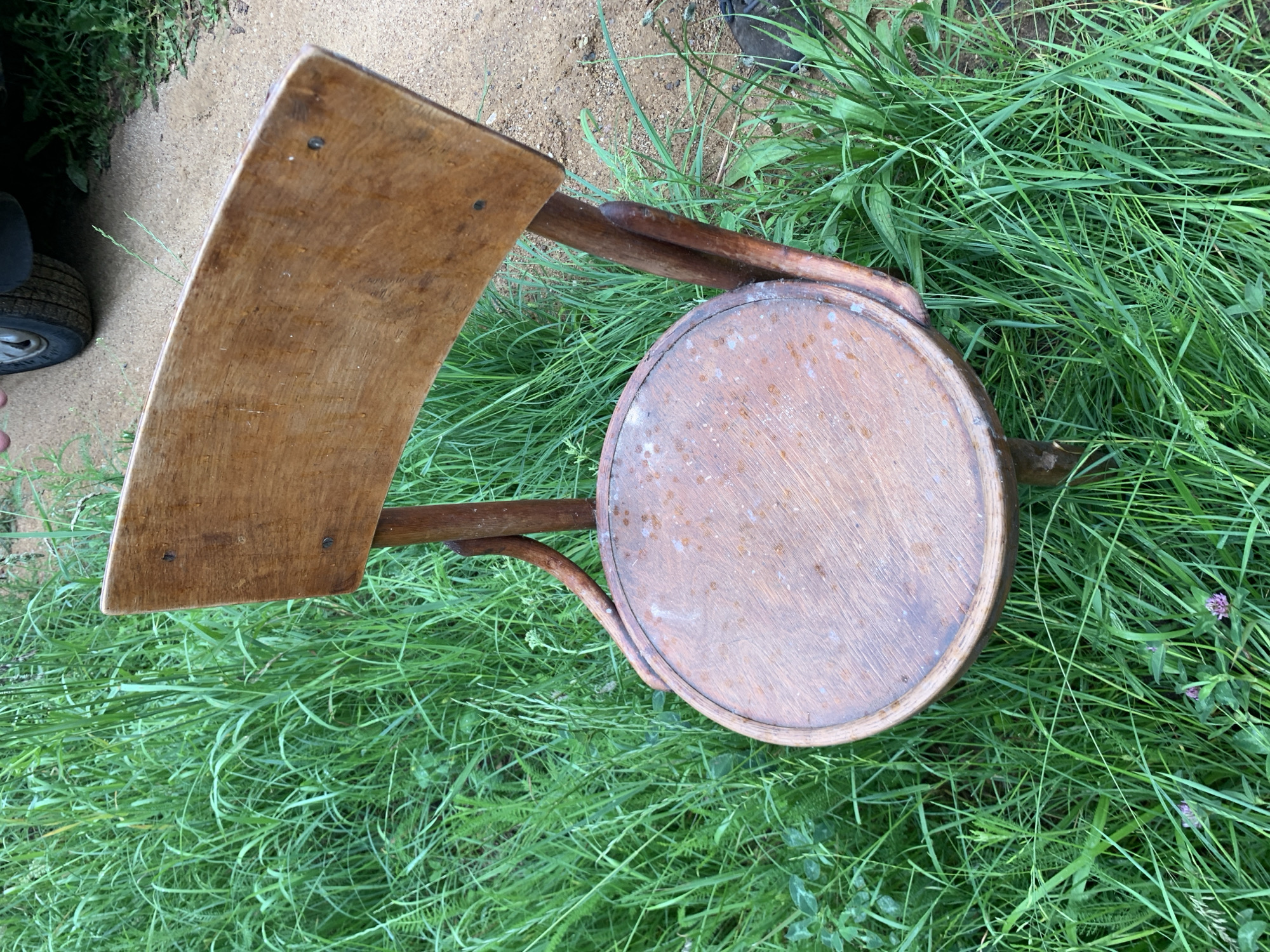 A weathered, wooden chair with a curved back and round seat sits on lush green grass and a dirt path. The chair appears to be old and shows signs of wear, with water stains and visible nail heads. The background features tall, dense grass and a sandy or gravelly path, suggesting an outdoor setting possibly in a rural or less-maintained area. A small portion of a car tire and fender are visible in the upper right corner, partially obscured by the chair and grass. The lighting suggests it is daytime, likely overcast given the lack of harsh shadows. The scene is devoid of human presence, focusing entirely on the static object within its natural environment. The location, Блеўчыцкі сельскі Савет, Belarus, indicates a village or rural administrative region.
