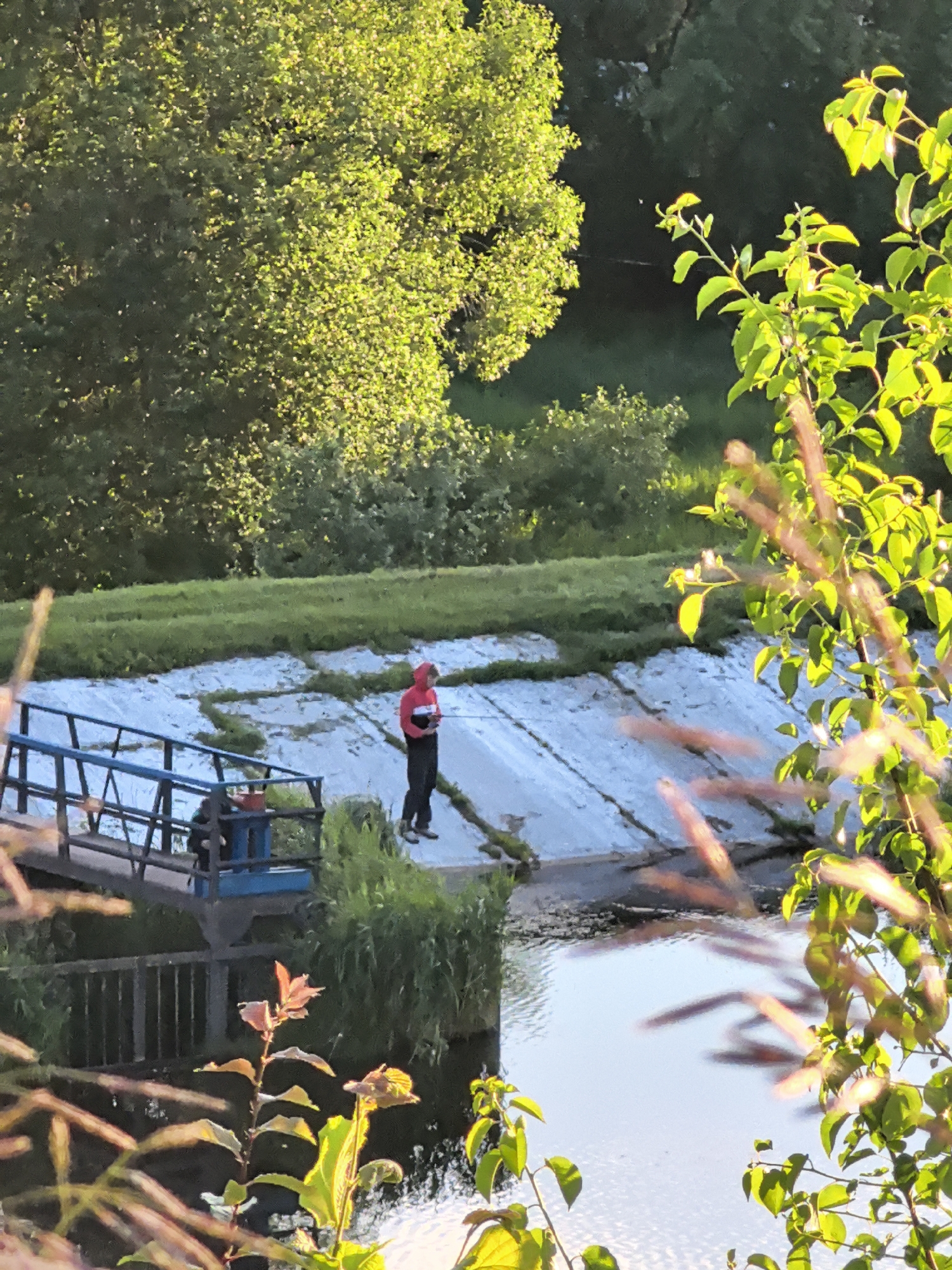 A person wearing a red hooded jacket and black pants stands on a concrete embankment, holding a rod with fishing line extending into the water. The person appears to be fishing. To the left, a blue metal walkway leads to a platform where another person is seated, with a red bucket visible. The embankment is covered in light-colored concrete slabs, with patches of grass growing between them. The scene is set in an outdoor environment with lush green trees and vegetation in the background and foreground. The water is calm and reflects the sky and surrounding foliage. The lighting suggests it is either early morning or late afternoon, with the sun casting a warm glow on the trees.