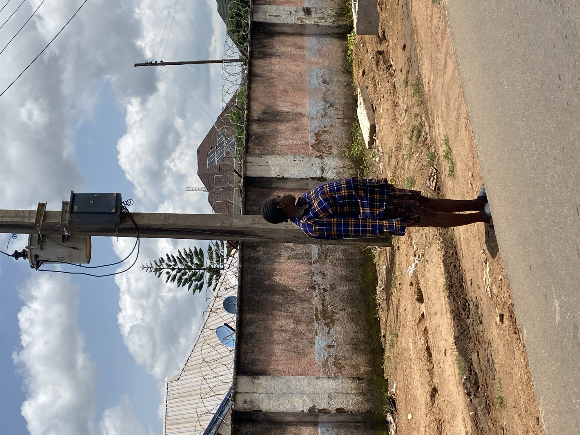 A medium shot captures a young Black woman standing on the side of a road in Gura Top, Nigeria, facing right and looking away from the camera. She is wearing a blue and yellow plaid long-sleeved shirt and patterned shorts. Her stance is neutral, with her feet shoulder-width apart. The woman stands near a weathered, pink and white painted concrete wall topped with barbed wire. Behind the wall, a rooftop with solar panels is visible, as well as a tall, conical evergreen tree. A utility pole with a transformer and electrical boxes is positioned behind and slightly to the left of the woman. The ground in front of the wall is a mixture of dirt, gravel, and sparse vegetation, with some debris scattered on the ground. The road in the foreground is paved asphalt. The sky is partly cloudy with white cumulus clouds against a blue background. The scene appears to be an outdoor, everyday urban environment.