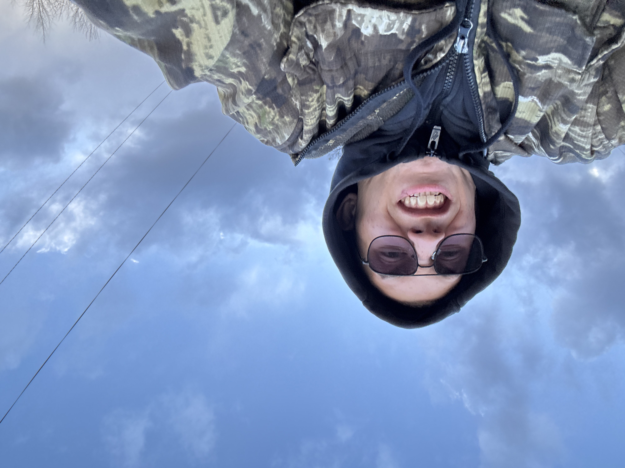 An inverted selfie captures a person, likely male, smiling broadly with teeth visible, looking directly at the camera. The person wears dark sunglasses, a black hooded sweatshirt with the hood up, and a camouflage-patterned jacket with a prominent zipper. The image appears to be taken from a low angle, looking upwards towards the subject.
The background primarily consists of a cloudy sky, displaying shades of light blue and grey, suggesting an overcast daytime setting. Several thin, dark lines, resembling power or utility lines, traverse the sky diagonally. In the top left corner of the original image (which would be the bottom left if viewed upright), the sparse branches of a bare tree are visible against the sky. The overall scene indicates an outdoor setting. No discernible text is visible in the image.