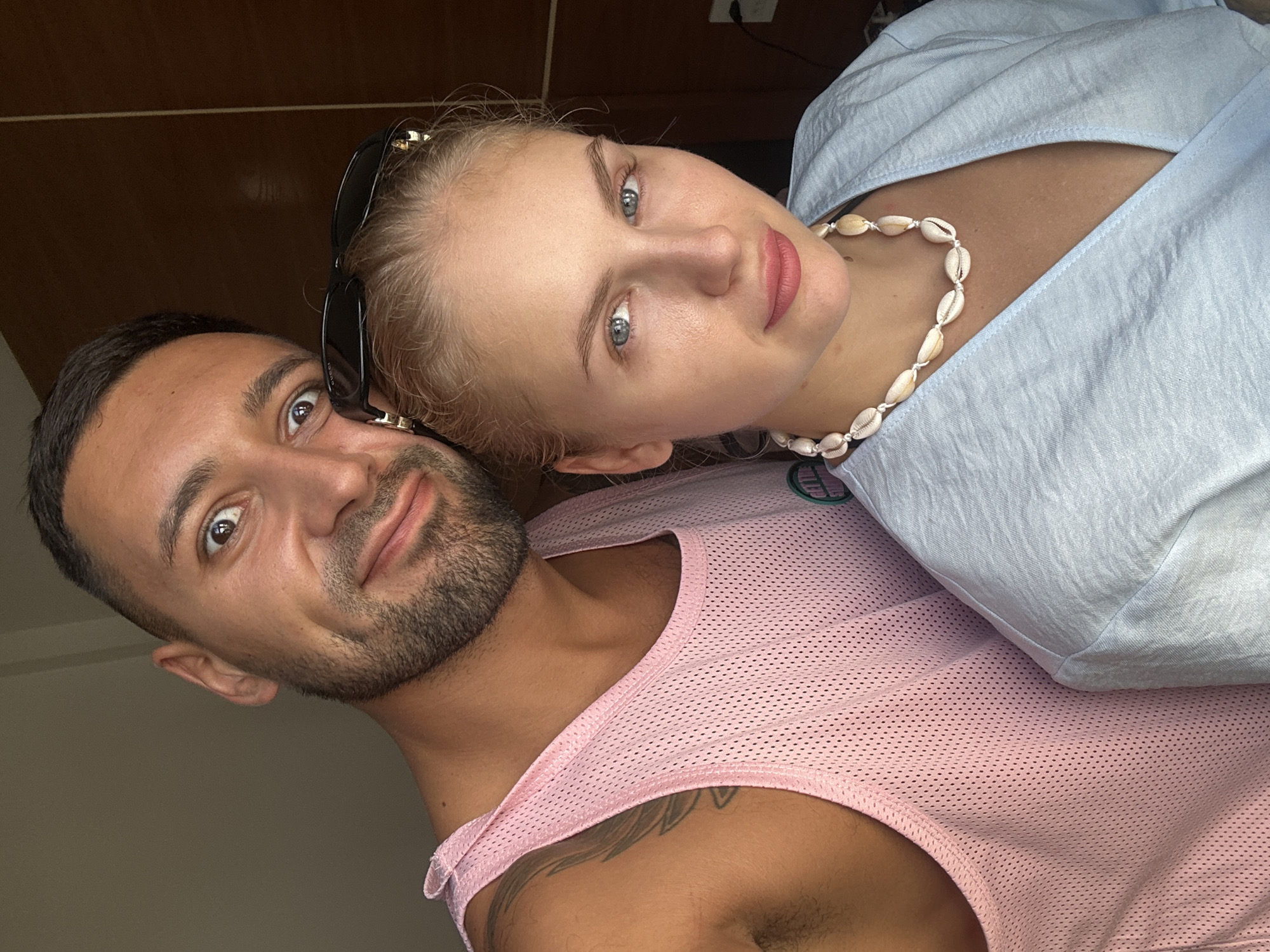 A man and a woman are posing for a selfie indoors, likely in Nha Trang Ward, Vietnam. The man, on the left, is wearing a pink mesh tank top and has short, dark hair and stubble. He is looking directly at the camera with a surprised expression. The woman, on the right, has blonde hair tied back and is wearing a light blue top and a shell necklace. Her sunglasses are perched on top of her head. The background appears to be a room with wooden paneling.