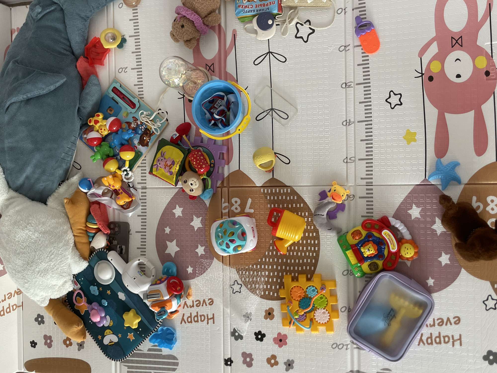 The image depicts a collection of baby toys scattered across a play mat. The play mat features a grid design with height measurements along the edges, along with repeating "Happy every" text and illustrations of stars and flowers.
The toys include a stuffed shark, a plush bear, a blue bucket with items inside, a musical toy, a set of gears, a plush giraffe, and other various play items. The toys are spread out across the mat, indicating a child's play area.
The OCR text on one of the toy packages is identified as Russian "АГНИЯ БАРТО ПЕРВЫЕ СТИХИ" (Agniya Barto First Poems). The mat also has "СХБ" as OCR text.
There is no indication of the location being in the city of Средний Постол, Russia. There are no people in the image.