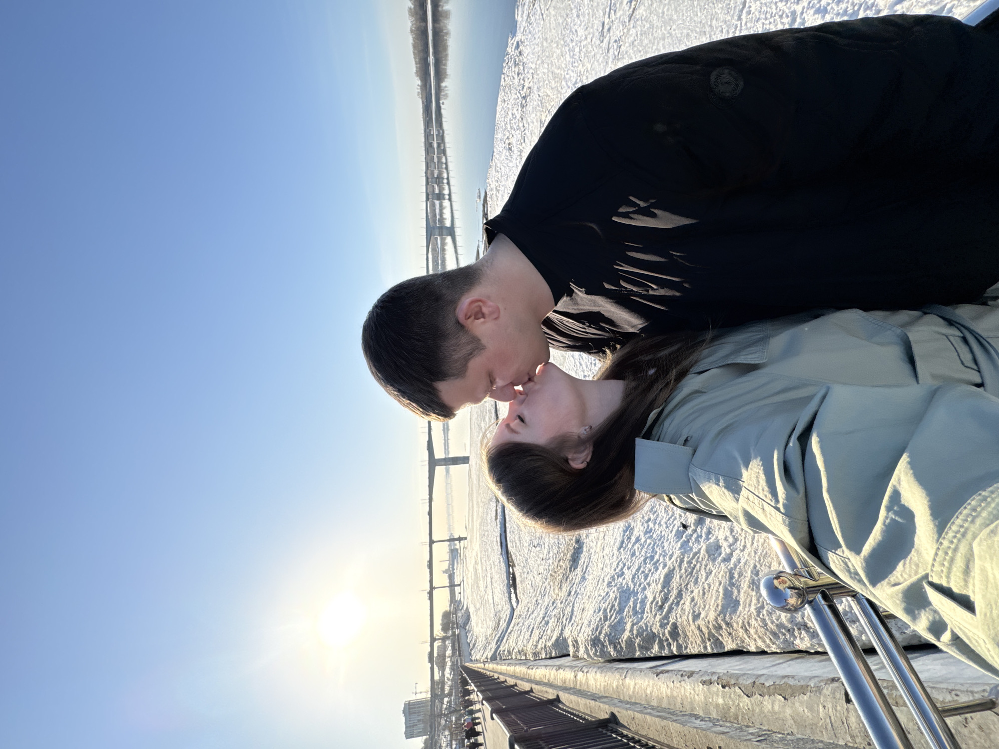 A medium outdoor shot depicts a couple kissing on a sunny day. The location is the embankment of the Kama River in the city of Perm, Russia. The couple is positioned in the foreground, with the man wearing a black jacket and the woman wearing a light green coat. A bridge stretches into the distance over a snowy landscape. The sky is clear and blue, with the sun positioned near the horizon. A metal railing is visible at the bottom of the frame. The image is oriented with the horizon at an angle.