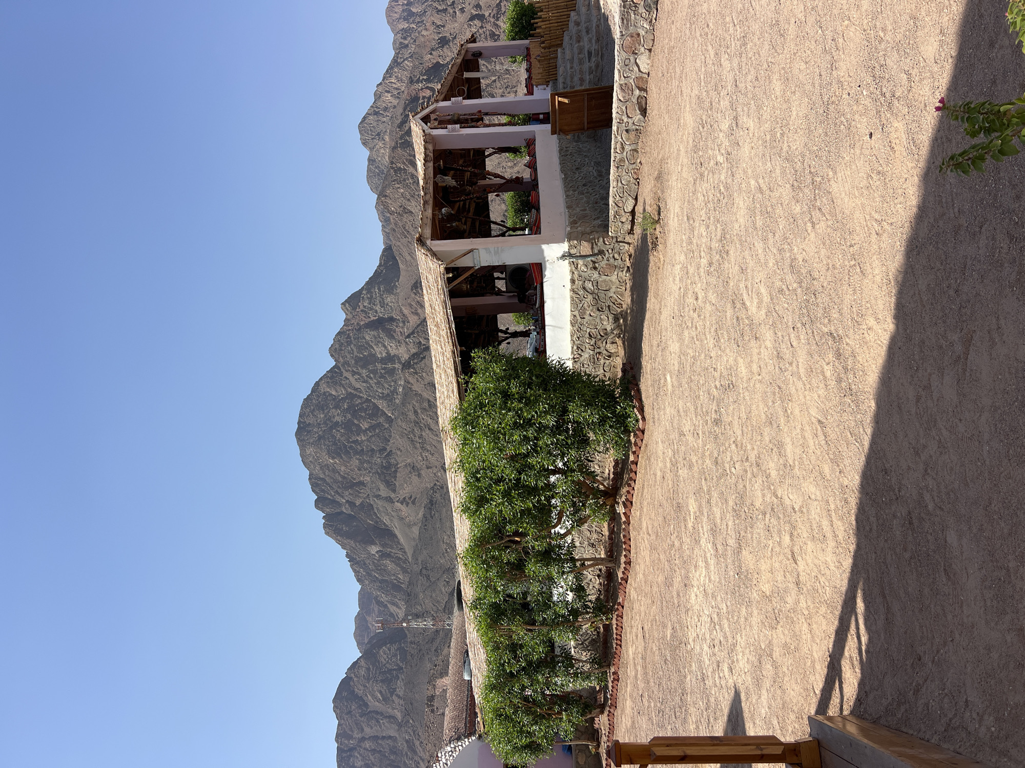 The image depicts an outdoor seating area at a establishment located in the city of Nueiba, Egypt. The establishment features a covered patio with a thatched roof, supported by pink-painted pillars. The patio is furnished with seating arrangements, including cushioned benches and cushions, suggesting a place for relaxation or dining. The exterior of the building is constructed with a stone base. In front of the patio, a neat row of green bushes are planted, bordered by a brick foundation. The ground is covered with fine gravel. In the background, rugged, mountainous terrain under a clear blue sky dominates the landscape. A telecommunications tower is visible on one of the distant mountains. A wooden fence and a staircase are also present to the right of the patio. The overall scene suggests a tranquil, arid environment common to the region.