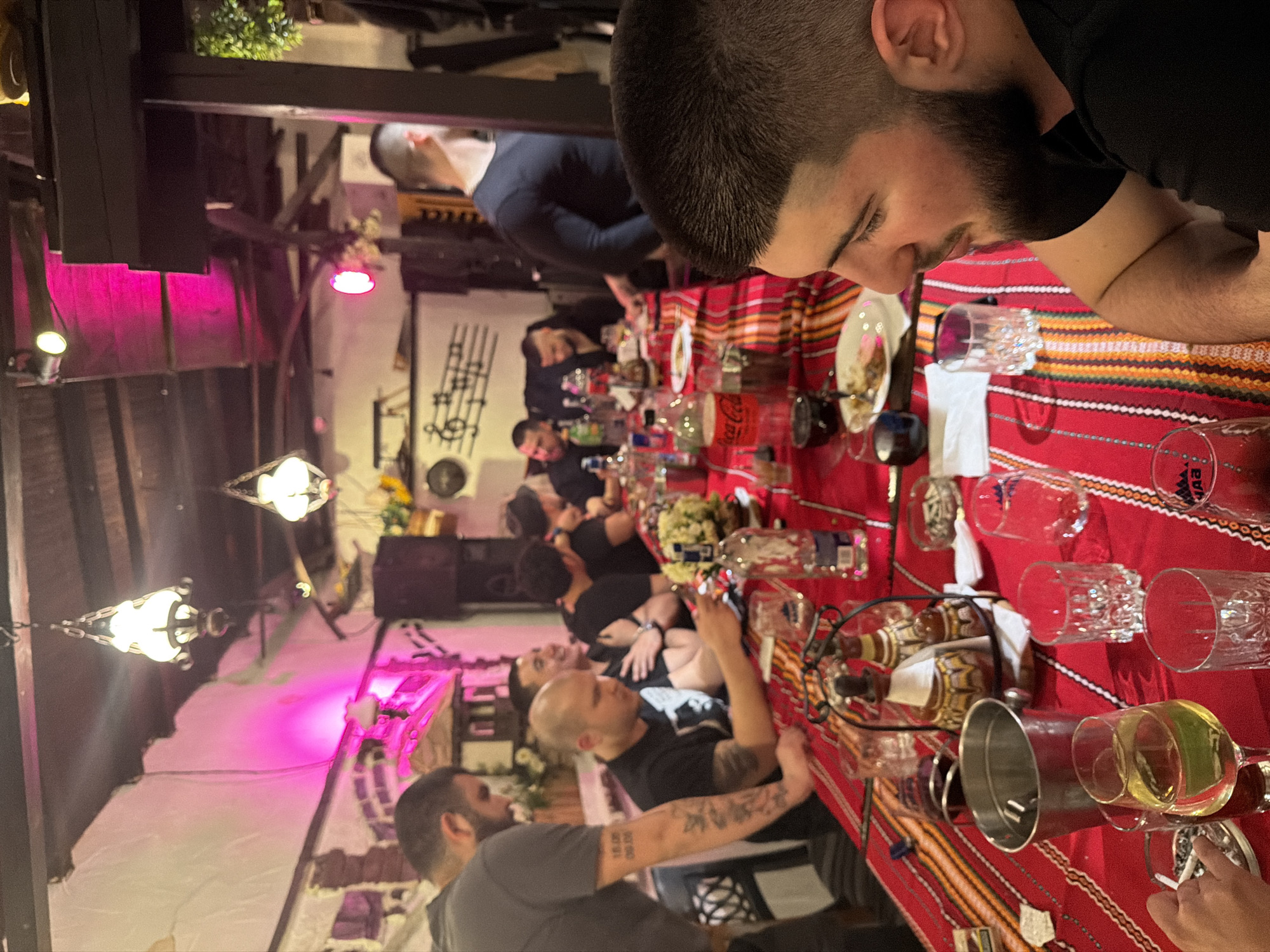 A long table covered with a red patterned cloth sits in a room with a wooden ceiling and dim lighting, possibly a restaurant in Varna, Bulgaria. Several people are seated around the table, engaged in conversation and dining. Numerous glasses and bottles are scattered across the table. A chandelier hangs from the ceiling. A man with dark hair and a beard is partially visible in the foreground.