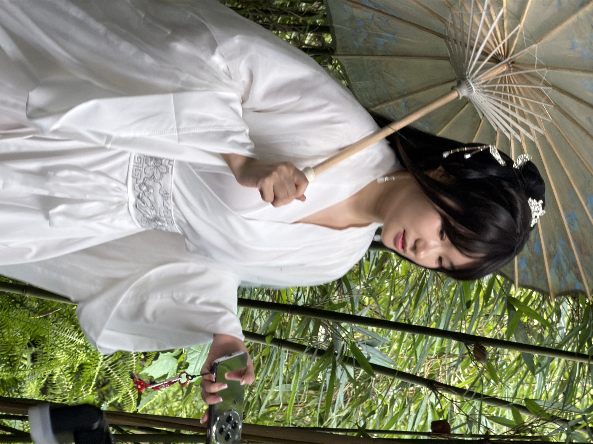 A woman in a white traditional Chinese garment stands in a bamboo forest. She holds a light-colored, patterned umbrella in her left hand, resting the handle on her shoulder. Her right hand holds a silver smartphone, which has a red keychain attached to it. The woman has black hair styled in a bun with decorative hairpins. She is wearing subtle makeup and a pearl earring. The bamboo stalks and lush green foliage form a dense backdrop. The scene appears to be outdoors, possibly in Yuzhong District, China, given the presence of bamboo and traditional attire.