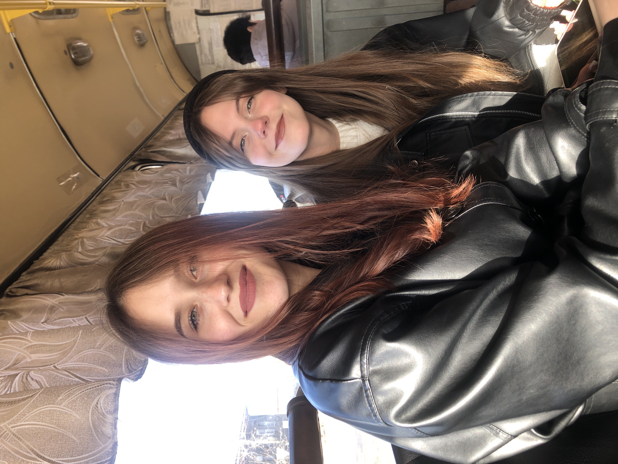 Two young adult females are seated inside a public transport vehicle, likely a bus, illuminated by bright daylight entering from a window on their left. Both subjects are looking towards the camera with slight smiles.
The female on the left has long, reddish-brown hair, light eyes, and faint freckles. She is wearing dark pink lipstick and a black faux leather jacket. Her shoulders and upper torso are visible.
The female on the right is positioned slightly behind and to the right of the first. She has long, light brown hair with bangs and is wearing a black headband. Her eyes are light, and she has a hint of redness on her nose. She is wearing nude lipstick and a black faux leather jacket over a white knit top. Her hands are visible in the lower right, holding a dark smartphone.
In the background, the back of a third individual's head and upper torso are visible, seated further back in the vehicle, wearing a light-colored hooded jacket.
The vehicle's interior features a curved, light tan ceiling with overhead light fixtures, yellow vertical grab poles on the right, and brown patterned curtains with a stylized leaf design. Through the window on the left, bare trees and a distant building are visible under bright daylight, suggesting an outdoor, possibly suburban or semi-rural, environment. The specific location within the city of Фоки, Russia, cannot be confirmed from the visual information presented.